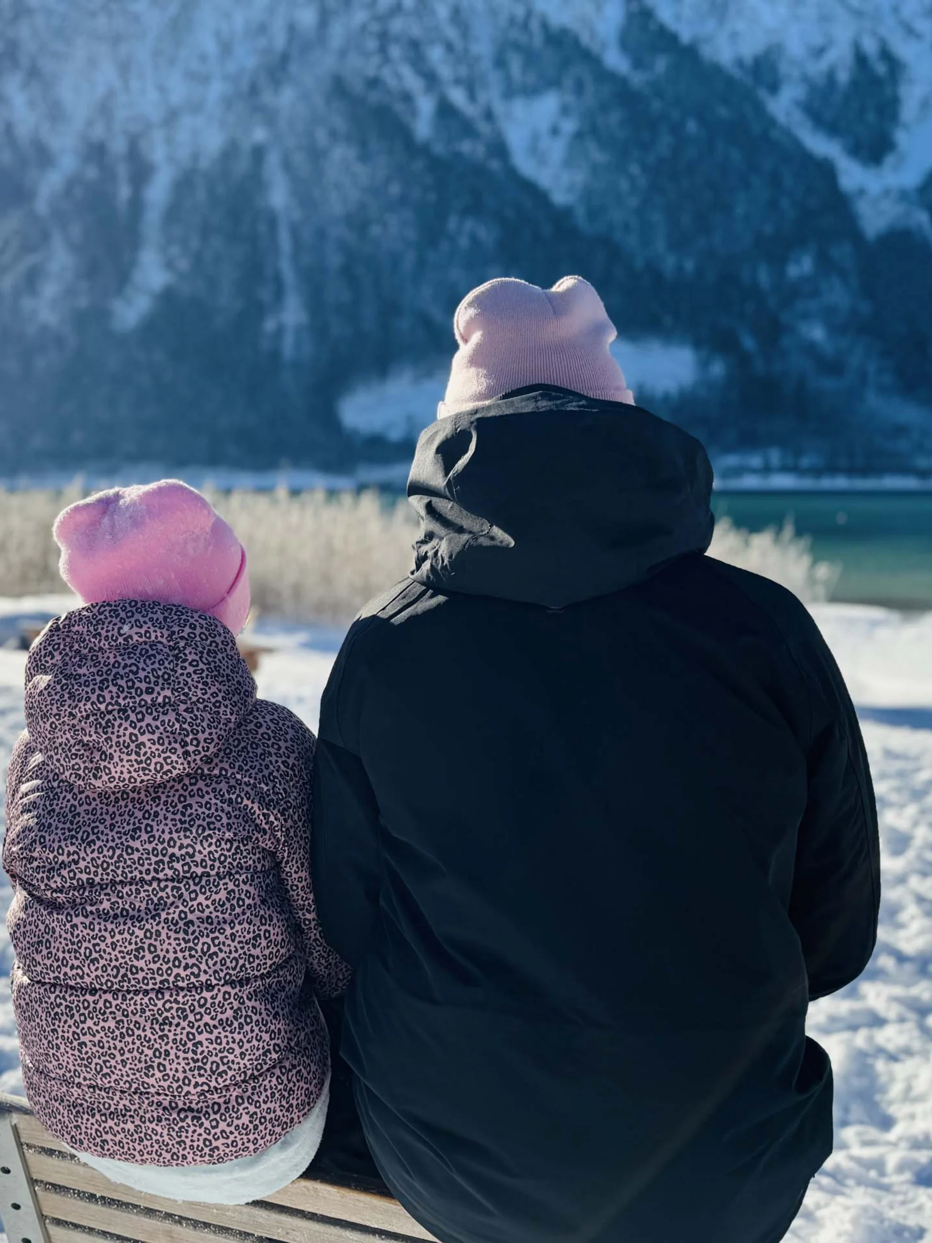 Welcome to our family hotel at Lake Achensee. Two people wearing hats sit on a bench in the snow facing snowy mountains