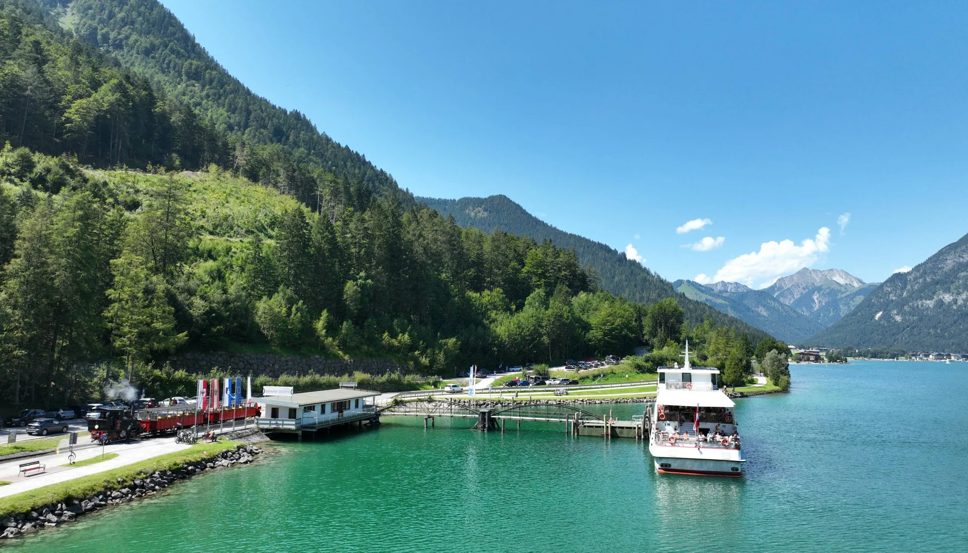 Grünes Bergseeufer mit Dampflock und Ausflugsboot bei Sonnenschein