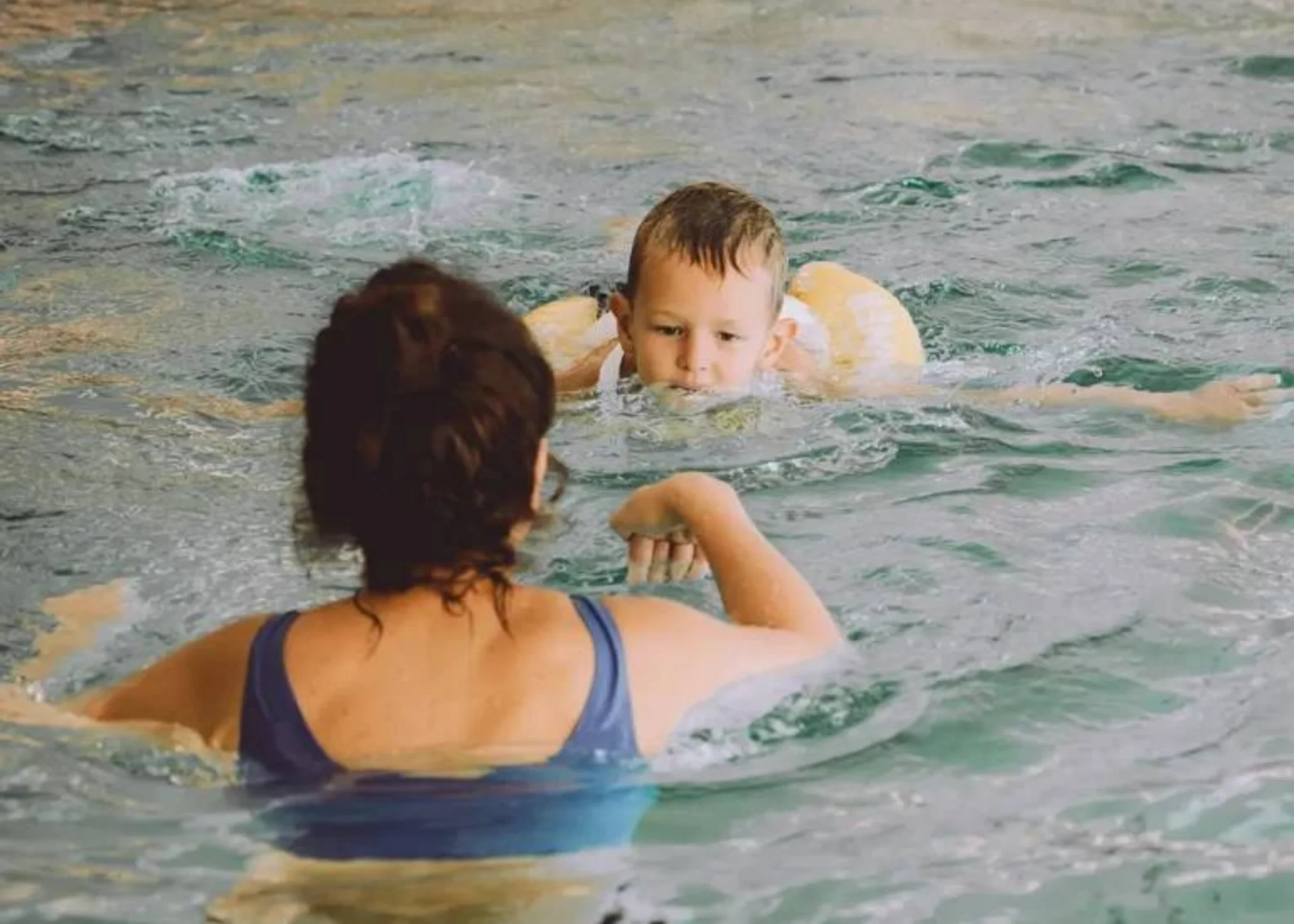 Frau unterrichtet Kind mit Schwimmhilfe im Hallenbad