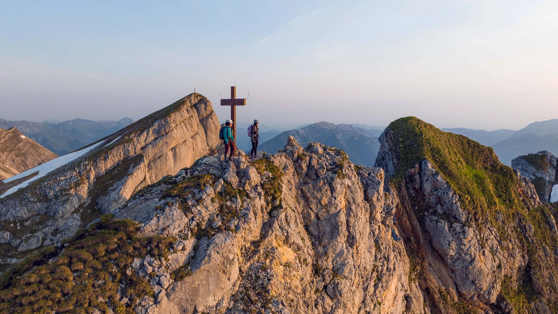 Zwei Wanderer auf felsigem Berggipfel neben Gipfelkreuz im Abendlicht