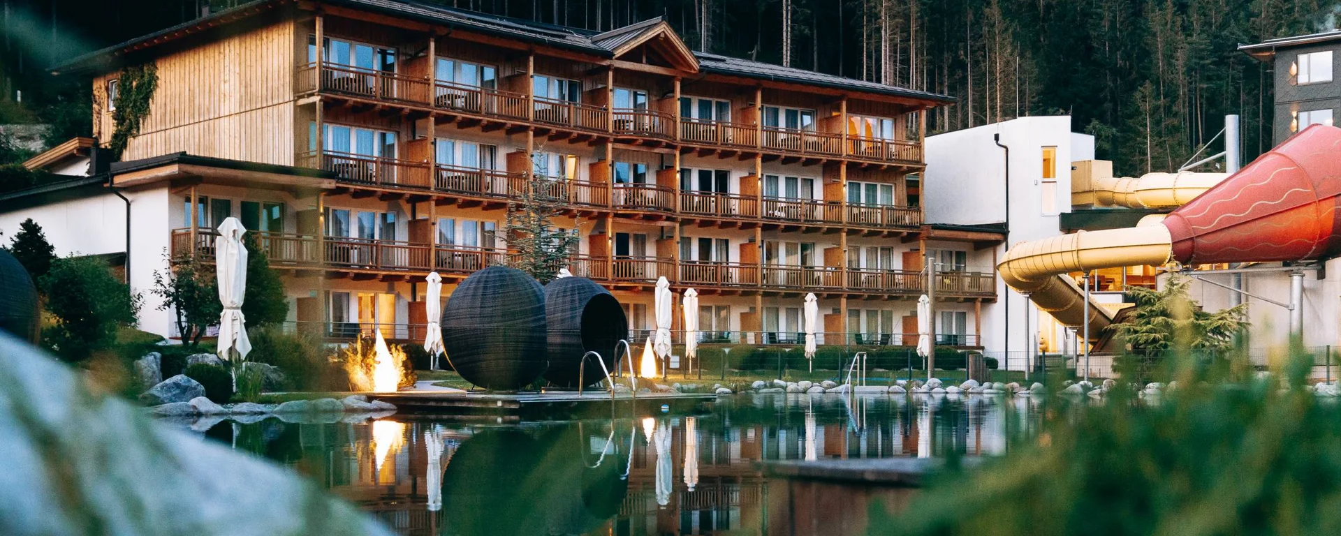 Hotel mit Holzfassade und Wasserrutsche neben einem See vor Waldkulisse
