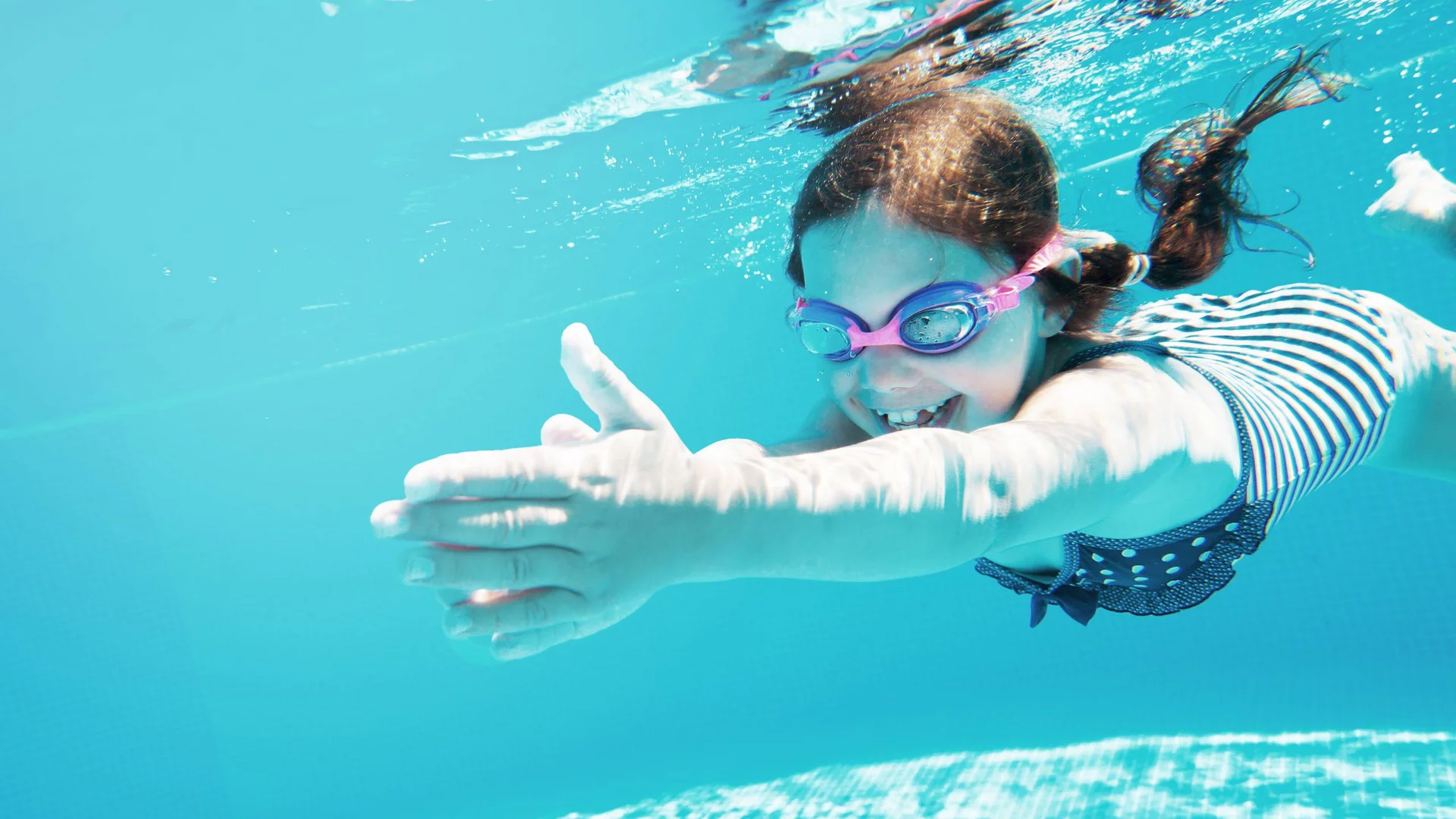 Lächendes Mädchen schwimmt unter Wasser im Pool mit Schwimmbrille und Badeanzug