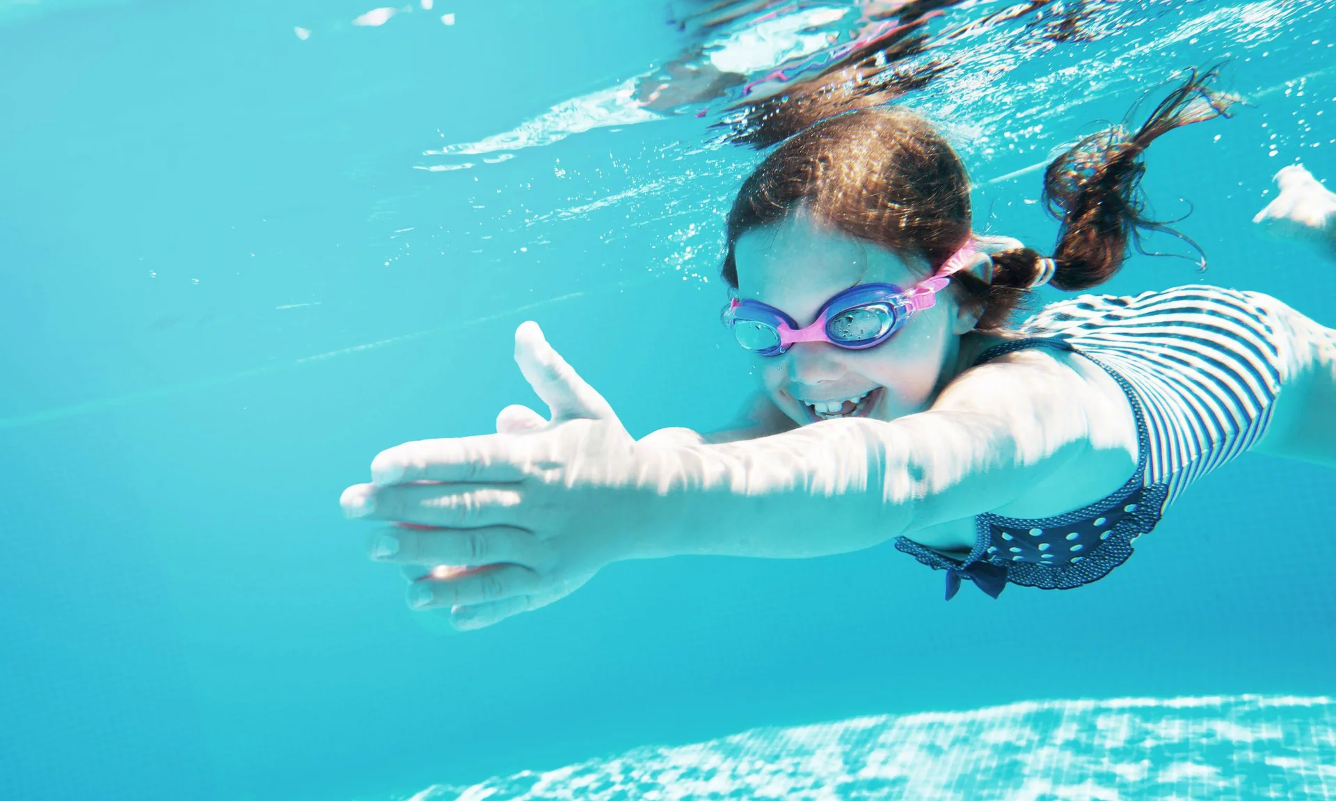 Lächendes Mädchen schwimmt unter Wasser im Pool mit Schwimmbrille und Badeanzug
