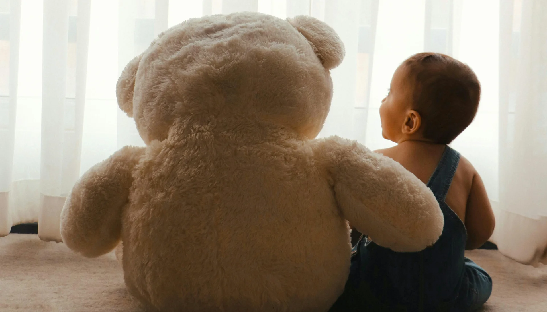 Baby sitting beside large teddy bear in front of white curtain