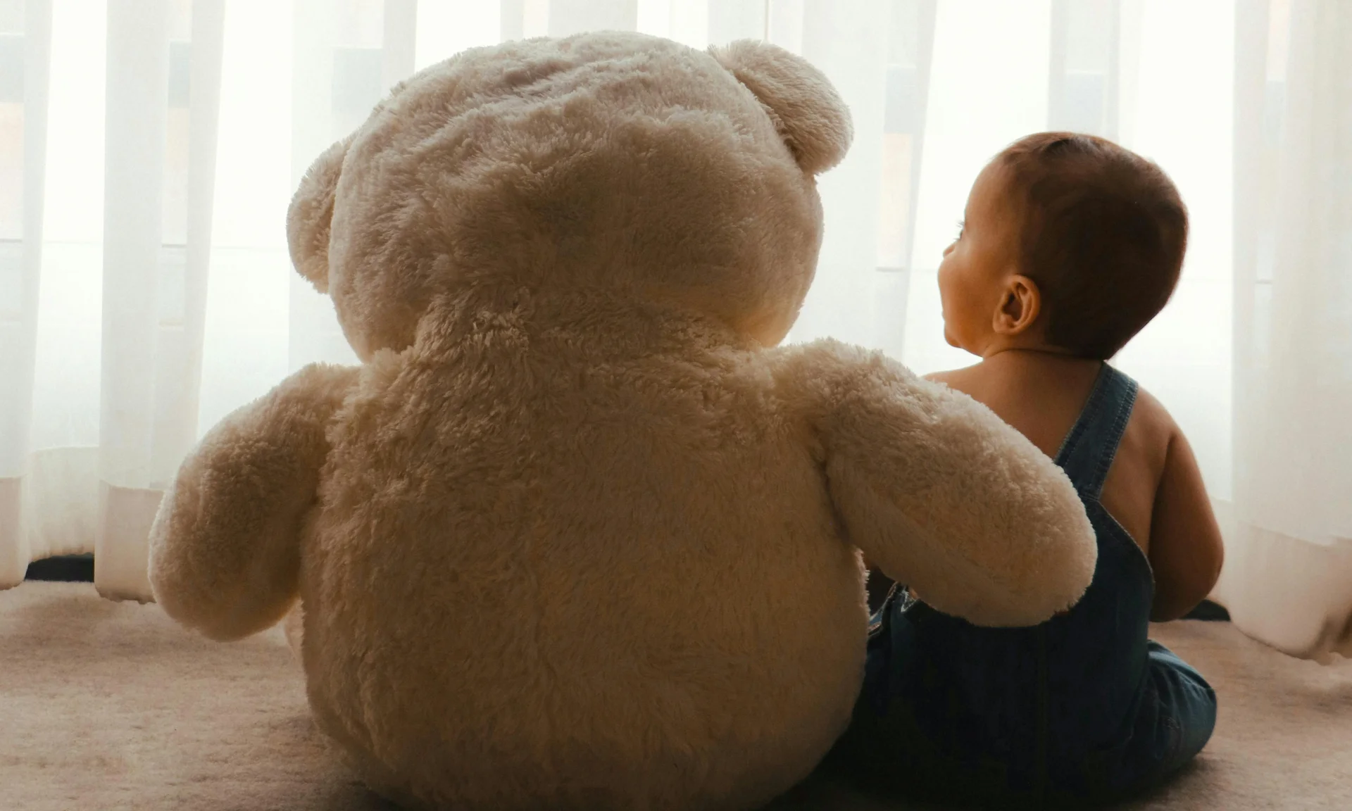 Baby sitzt neben großem Plüschbär vor weißem Vorhang