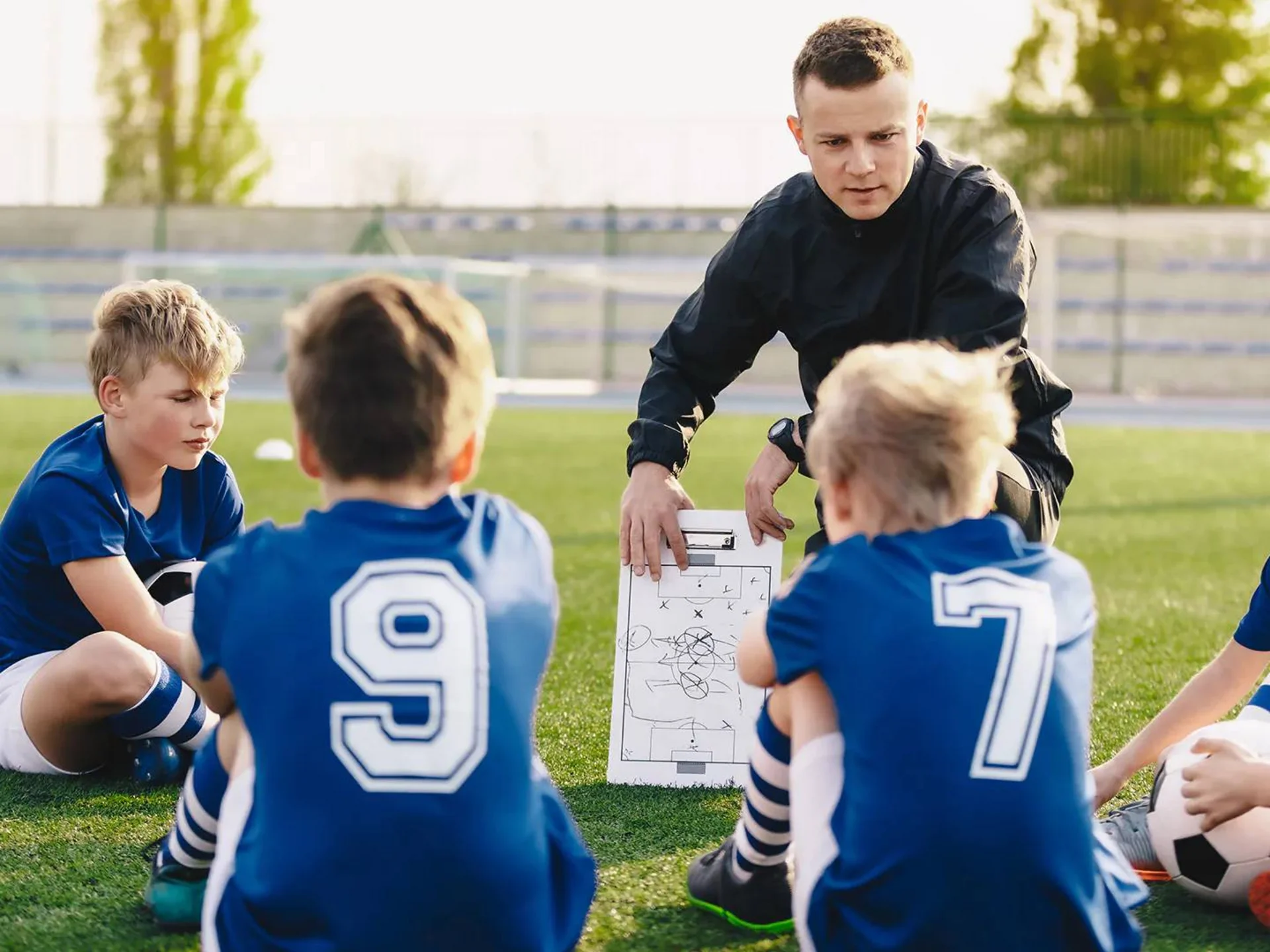Trainer erklärt Kindern Spieltaktiken mit Taktiktafel auf Fußballfeld