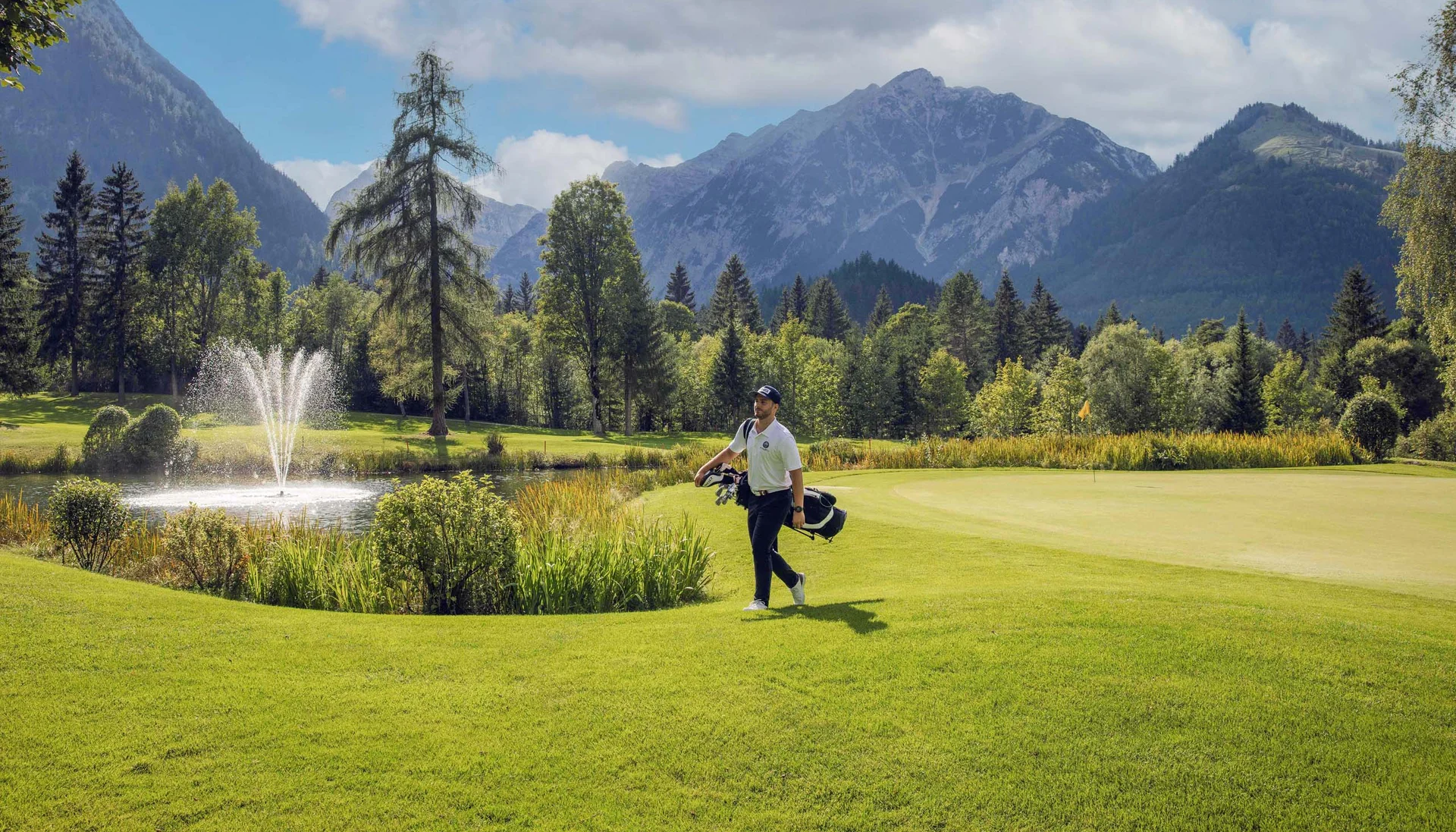 Golfer geht auf einem Rasenplatz mit Bergen und Springbrunnen im Hintergrund