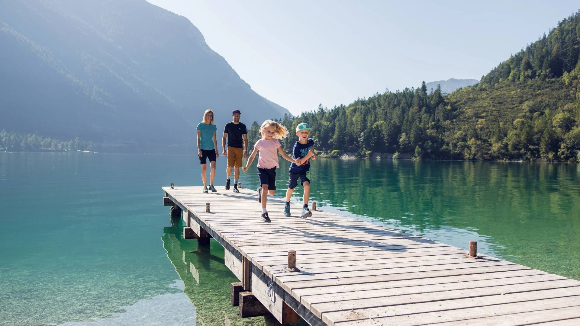 Familie mit zwei Kindern läuft auf einem Steg über einem Bergsee bei Sonnenschein