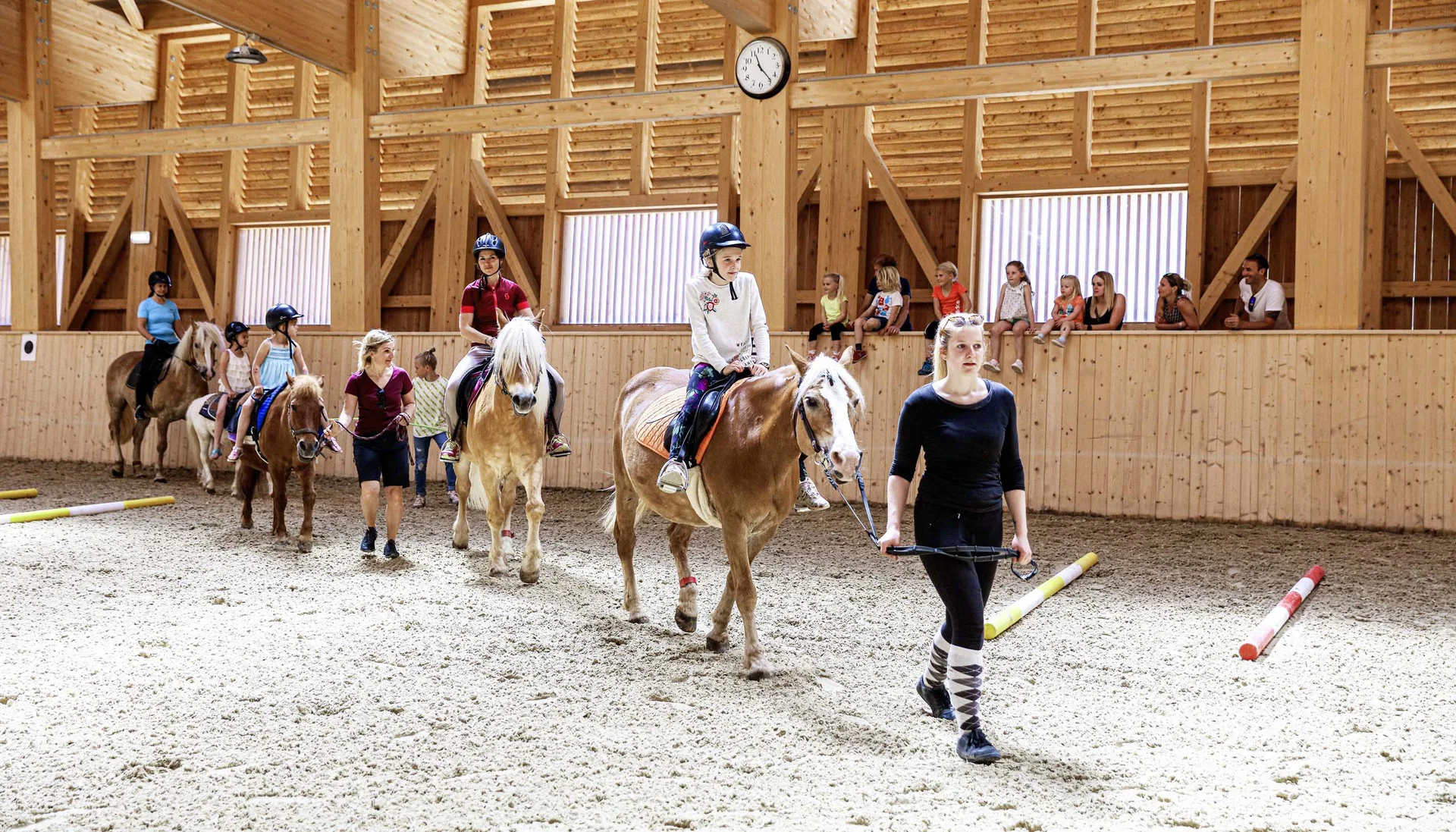 Kinder beim Reitunterricht in einer hellen Holzhalle, geführt von Erwachsenen