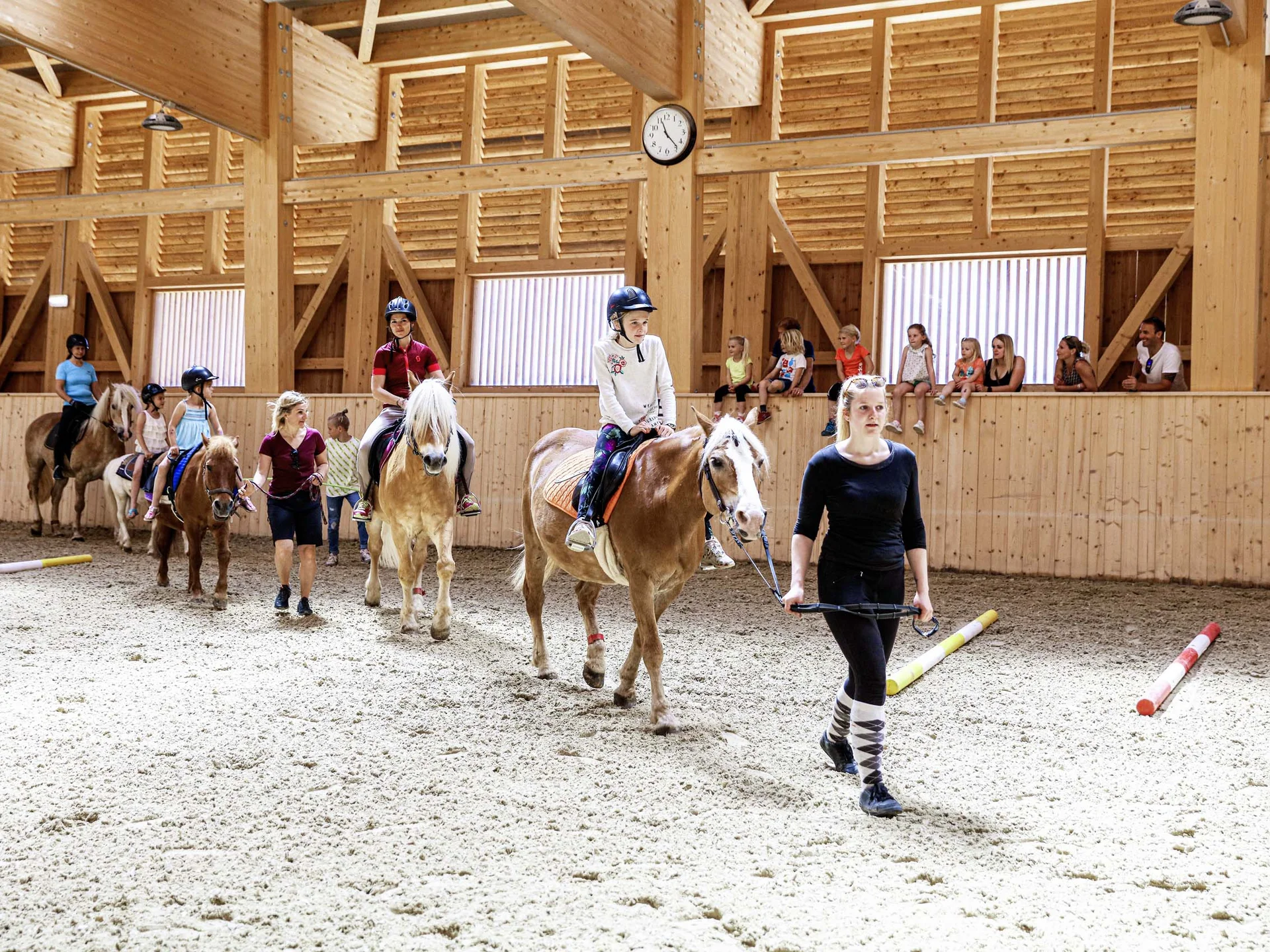 Kinder beim Reitunterricht in einer hellen Holzhalle, geführt von Erwachsenen