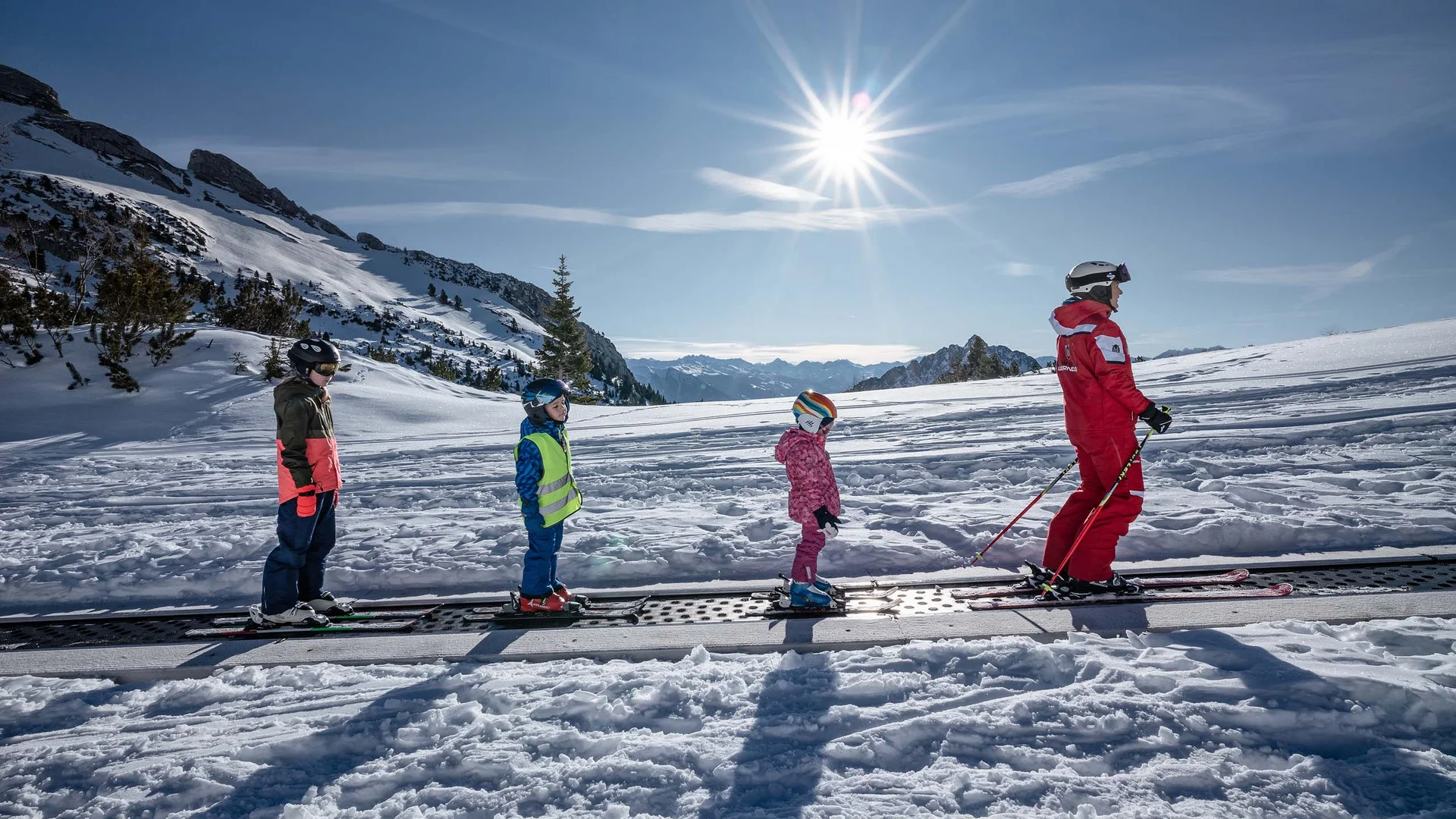 Skilehrer mit drei Kindern auf einem Förderband im verschneiten Skigebiet bei Sonnenschein