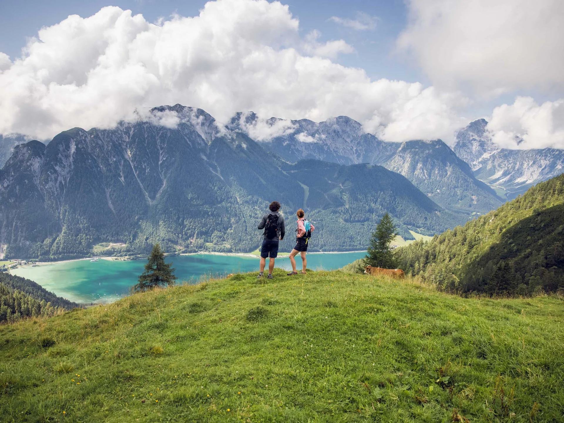 Zwei Wanderer stehen auf einer Wiese mit Berg- und Seeblick