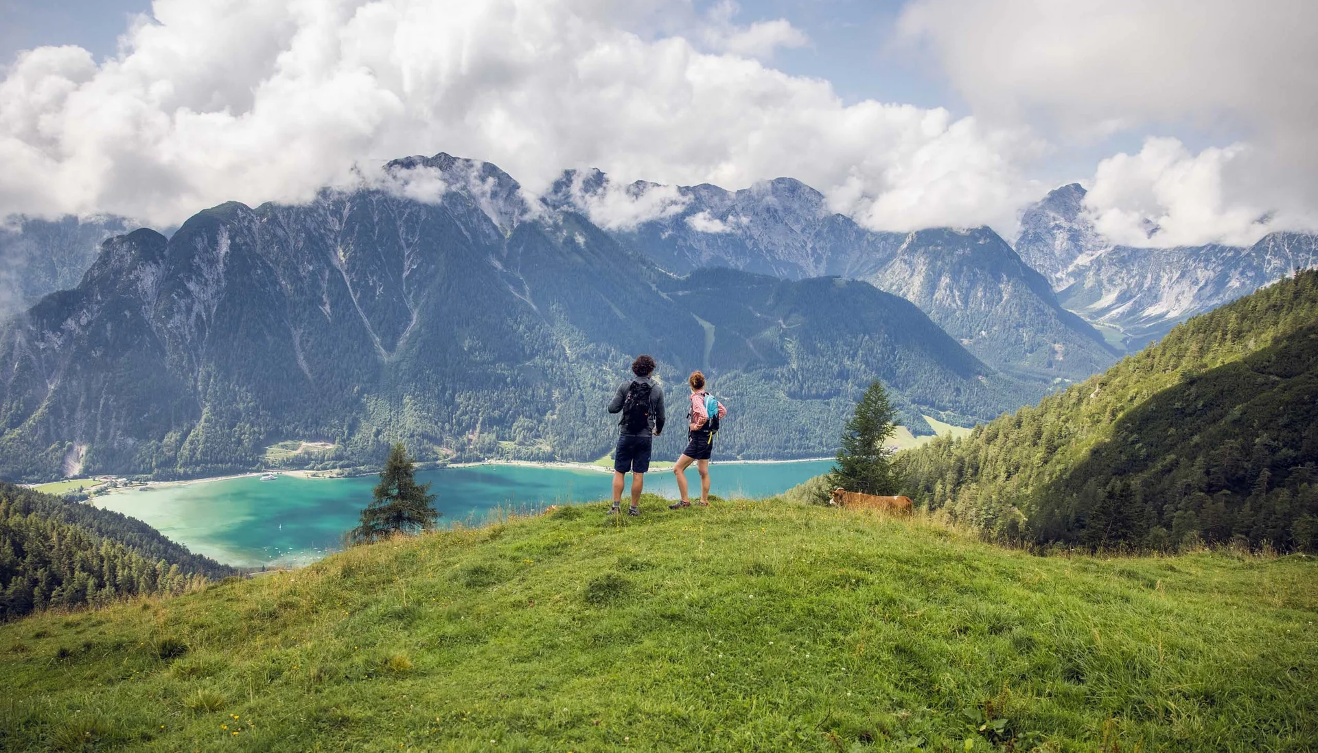 Zwei Wanderer stehen auf einer Wiese mit Berg- und Seeblick