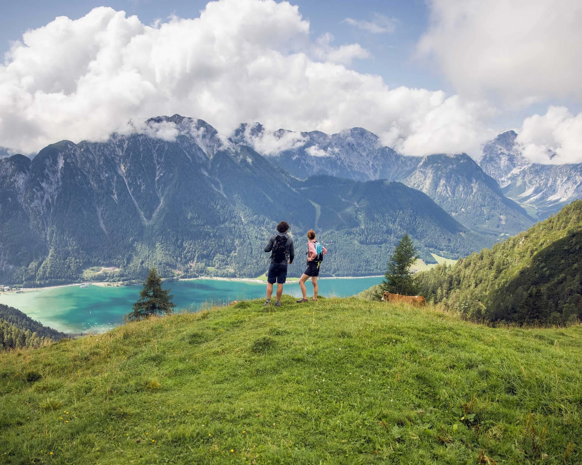 Zwei Wanderer stehen auf einer Wiese mit Berg- und Seeblick