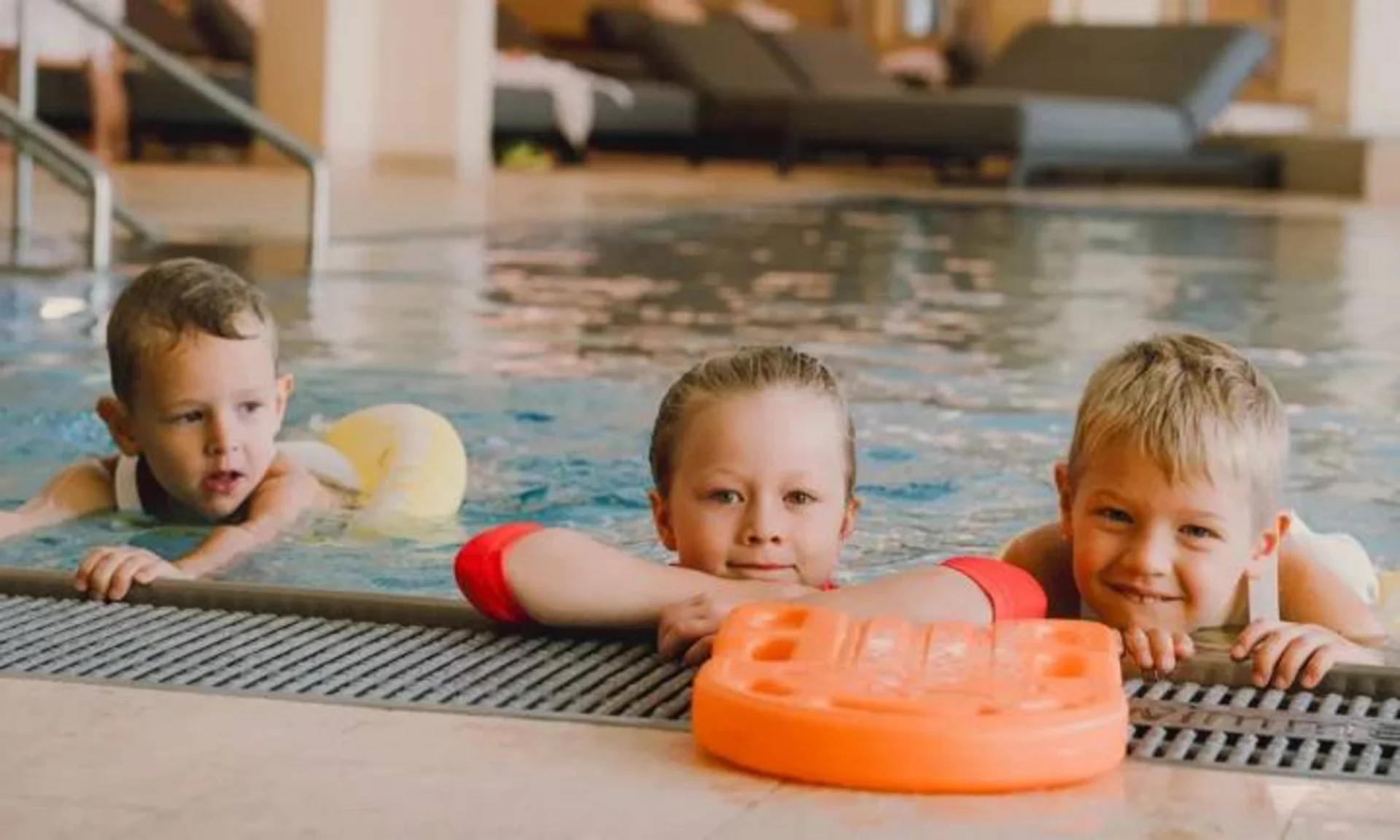 Three children in indoor pool holding onto pool edge with floaties