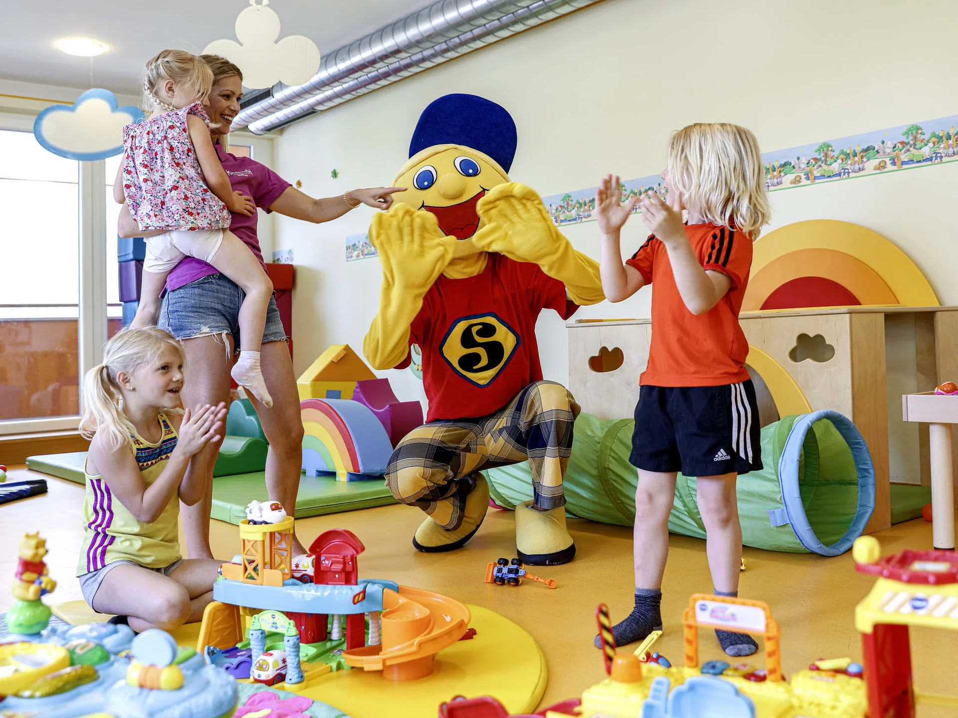 Children playing and clapping with a person in a colorful mascot costume in a playroom