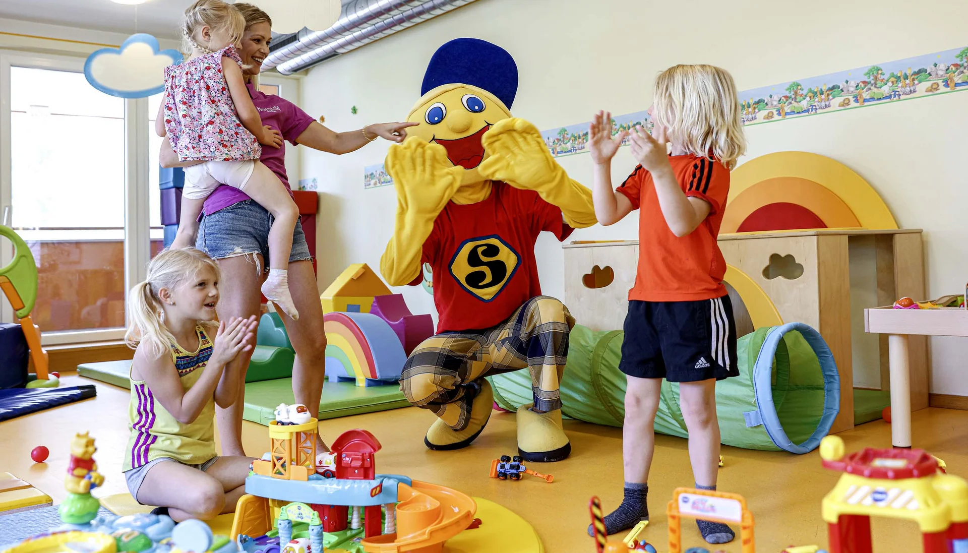 Kinder spielen und klatschen mit einer Person in einem bunten Maskottchenkostüm in einem Spielzimmer