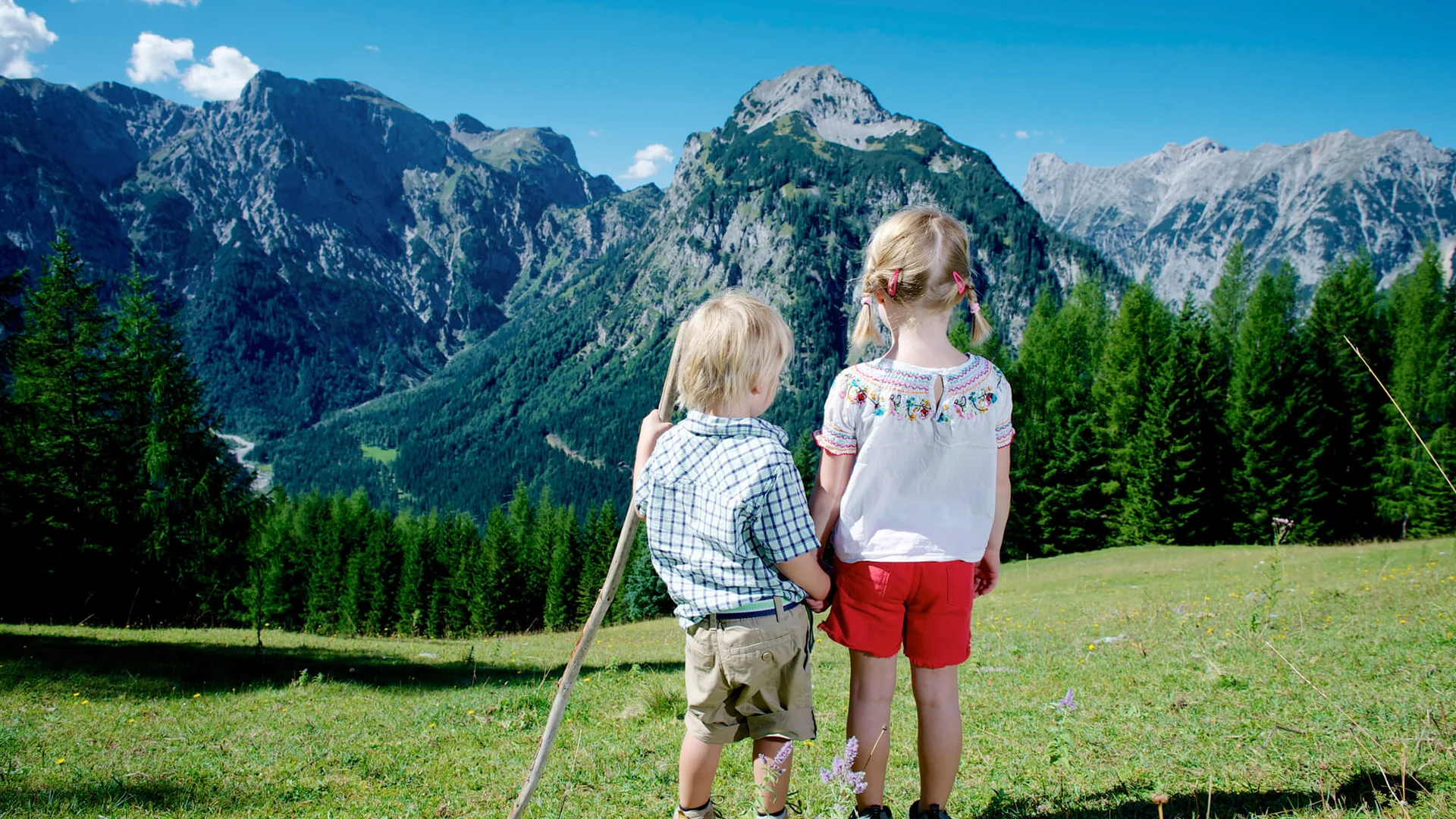 Kinder in den Alpen blicken auf Berge und Wälder unter blauem Himmel