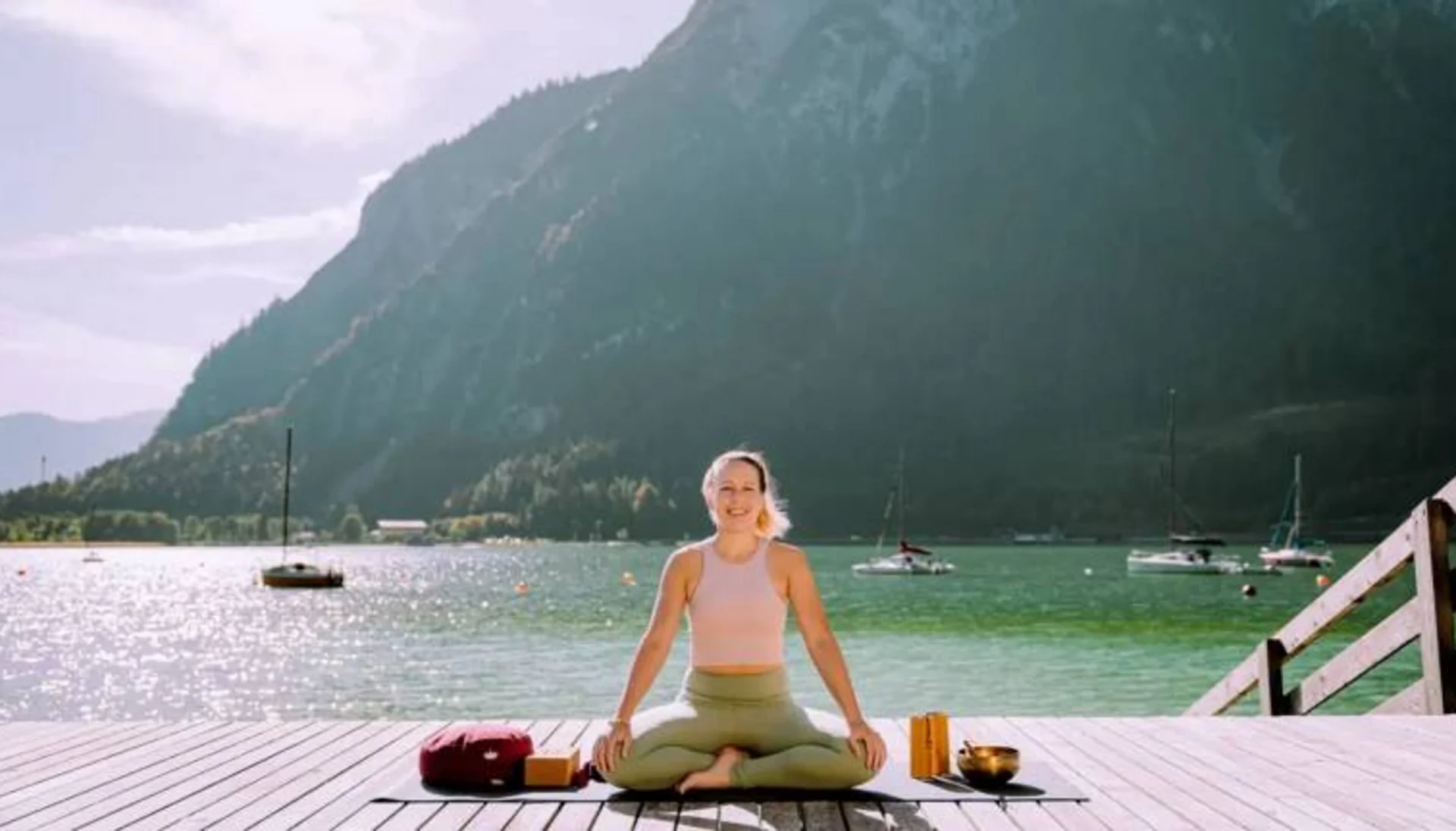 Frau macht Yoga auf Steg am See mit Bergen und Booten im Hintergrund
