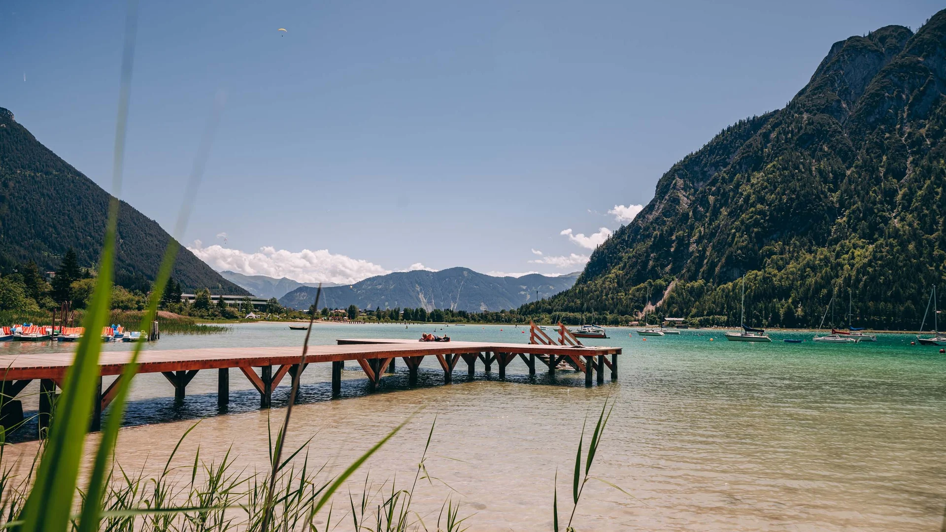 Holzsteg am klaren Bergsee mit Segelbooten und bewaldeten Bergen im Hintergrund