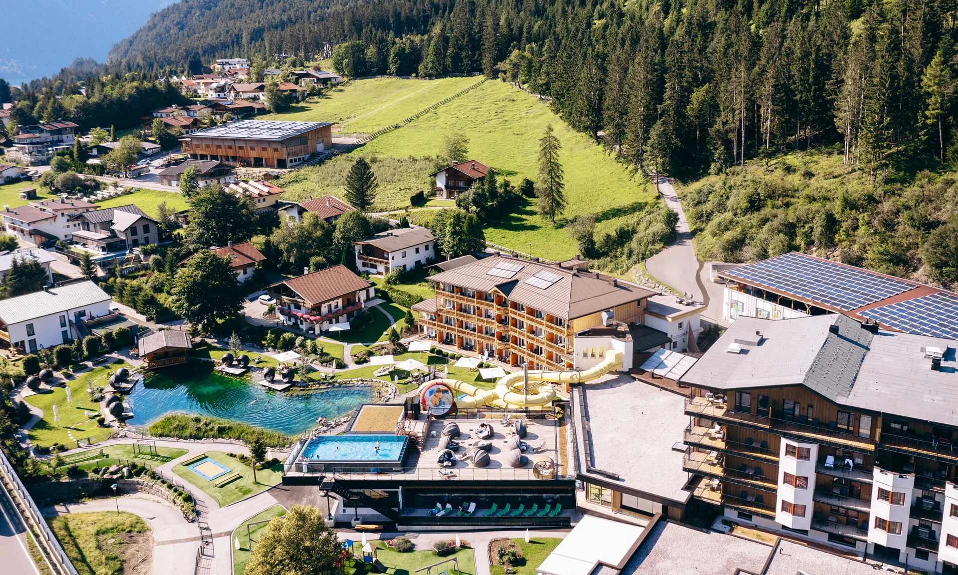 Luftaufnahme eines Resorts mit Swimmingpools und Wasserrutsche in grüner Alpenlandschaft