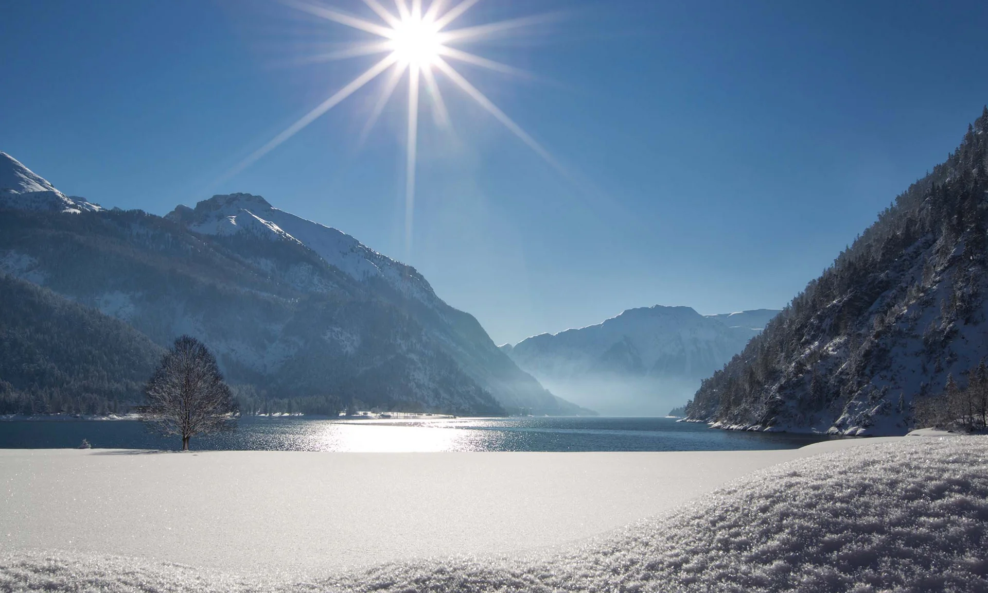 Schneebedeckter Bergsee bei Sonnenschein mit Bergen und blauem Himmel