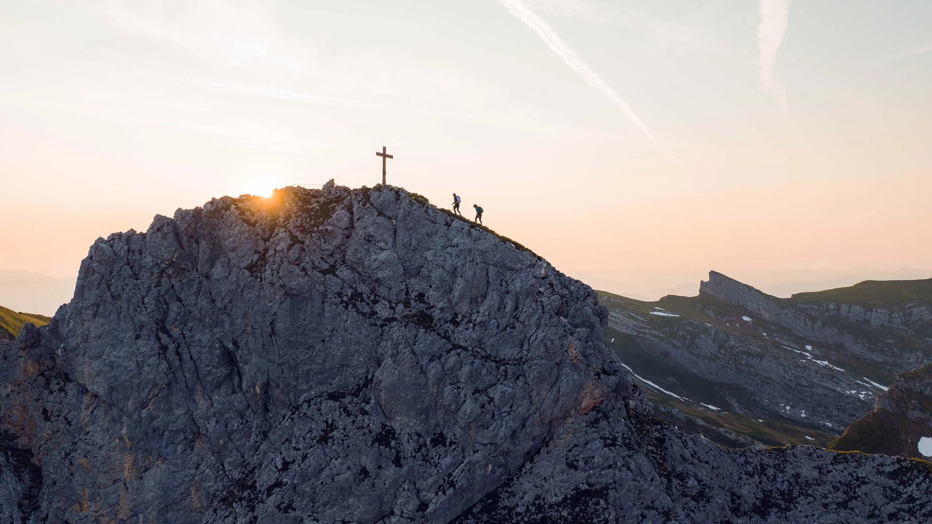 Zwei Wanderer steigen bei Sonnenuntergang auf einen Felsen mit Gipfelkreuz