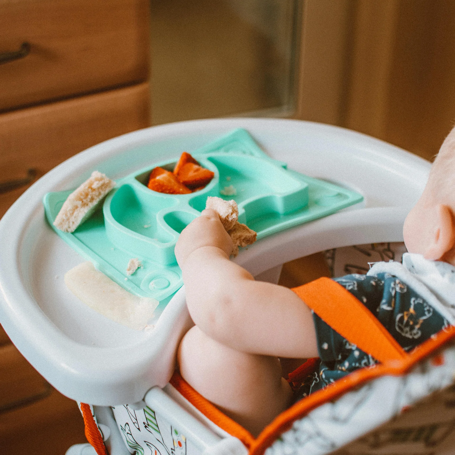 Baby in high chair eating bread and strawberries from a blue plate