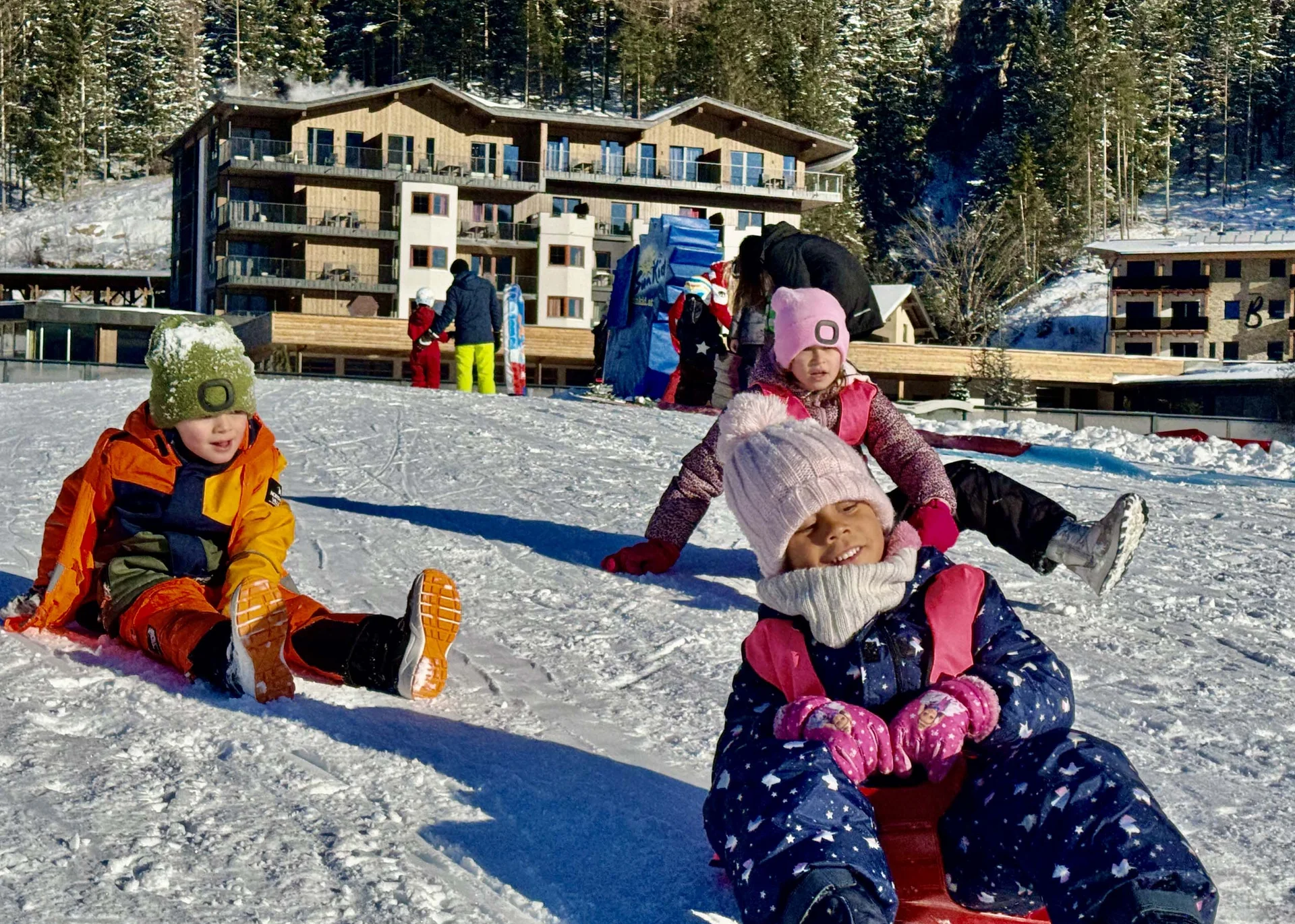 Ihr Familienhotel in Österreich mit All-inclusive-Angebot Kinder rodeln auf einem verschneiten Hügel vor einem Holzgebäude im Winter