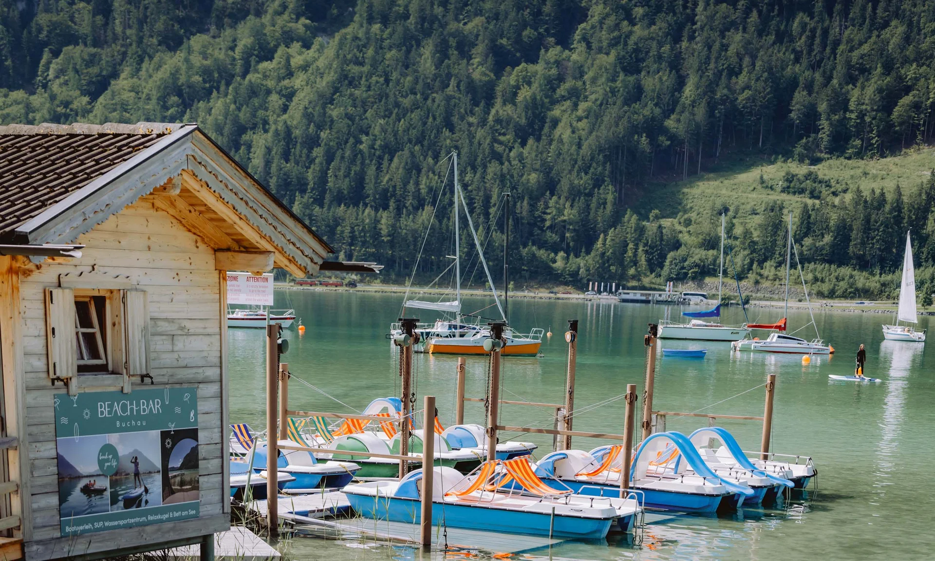 Ruderboote und Segelboote an einem Steg vor bewaldeten Bergen an einem See