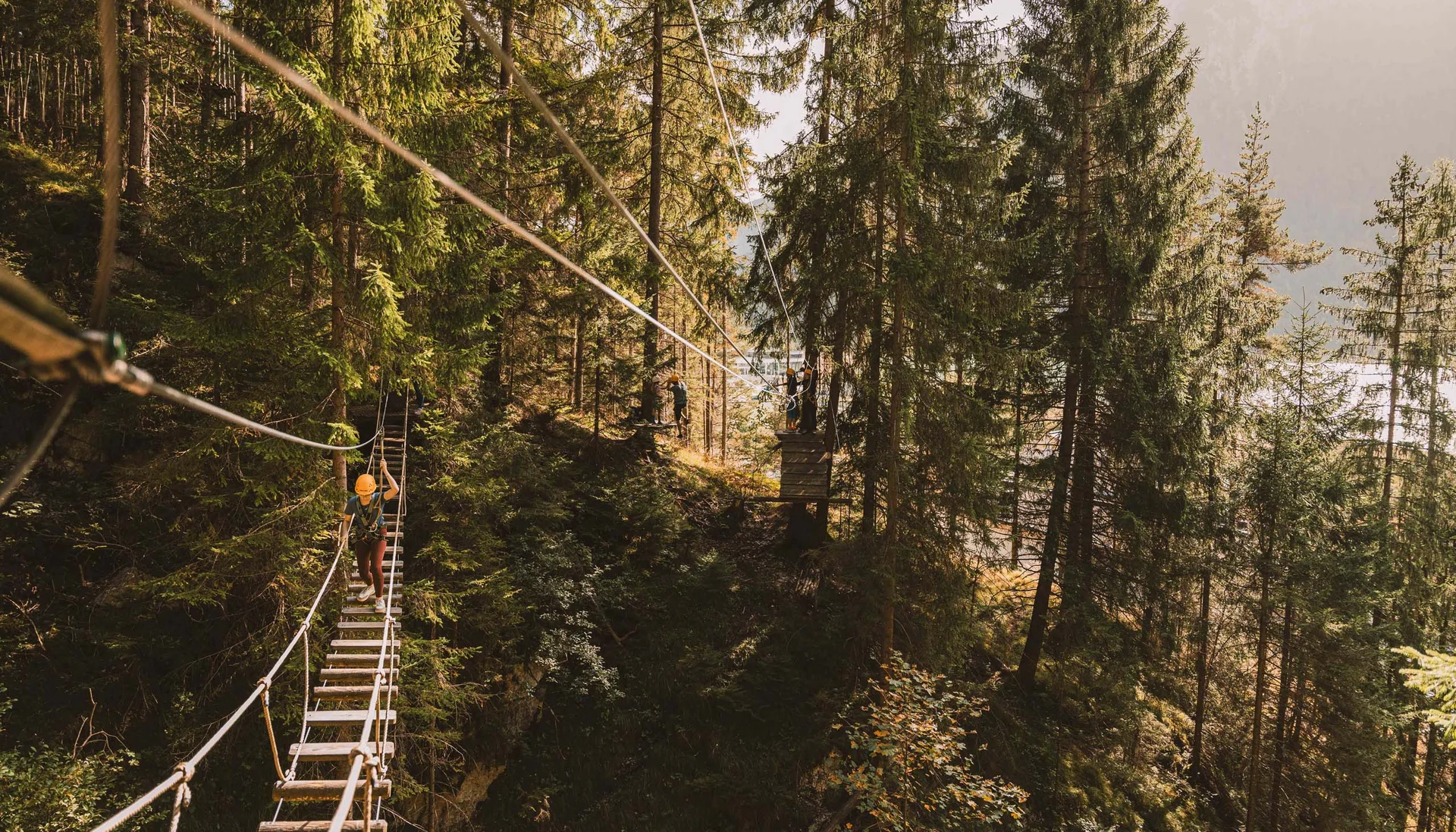 Person auf Hängebrücke in einem Kletterpark im Wald mit anderen Menschen im Hintergrund