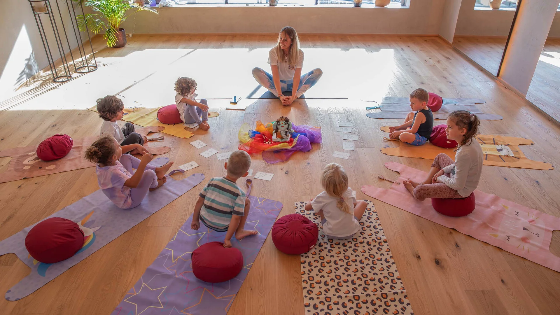 Kinder und Lehrerin sitzen im Kreis auf Yogamatten in einem hellen Raum