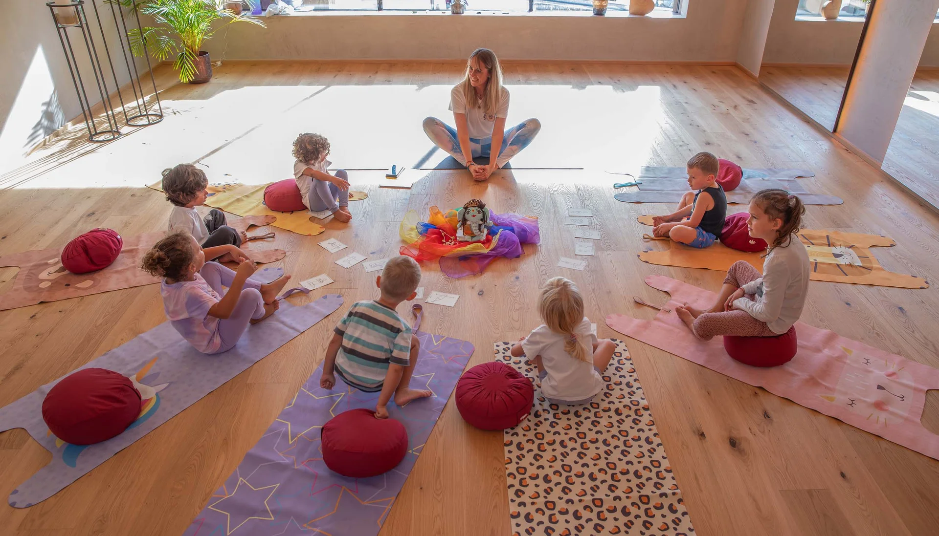 Kinder und Lehrerin sitzen im Kreis auf Yogamatten in einem hellen Raum