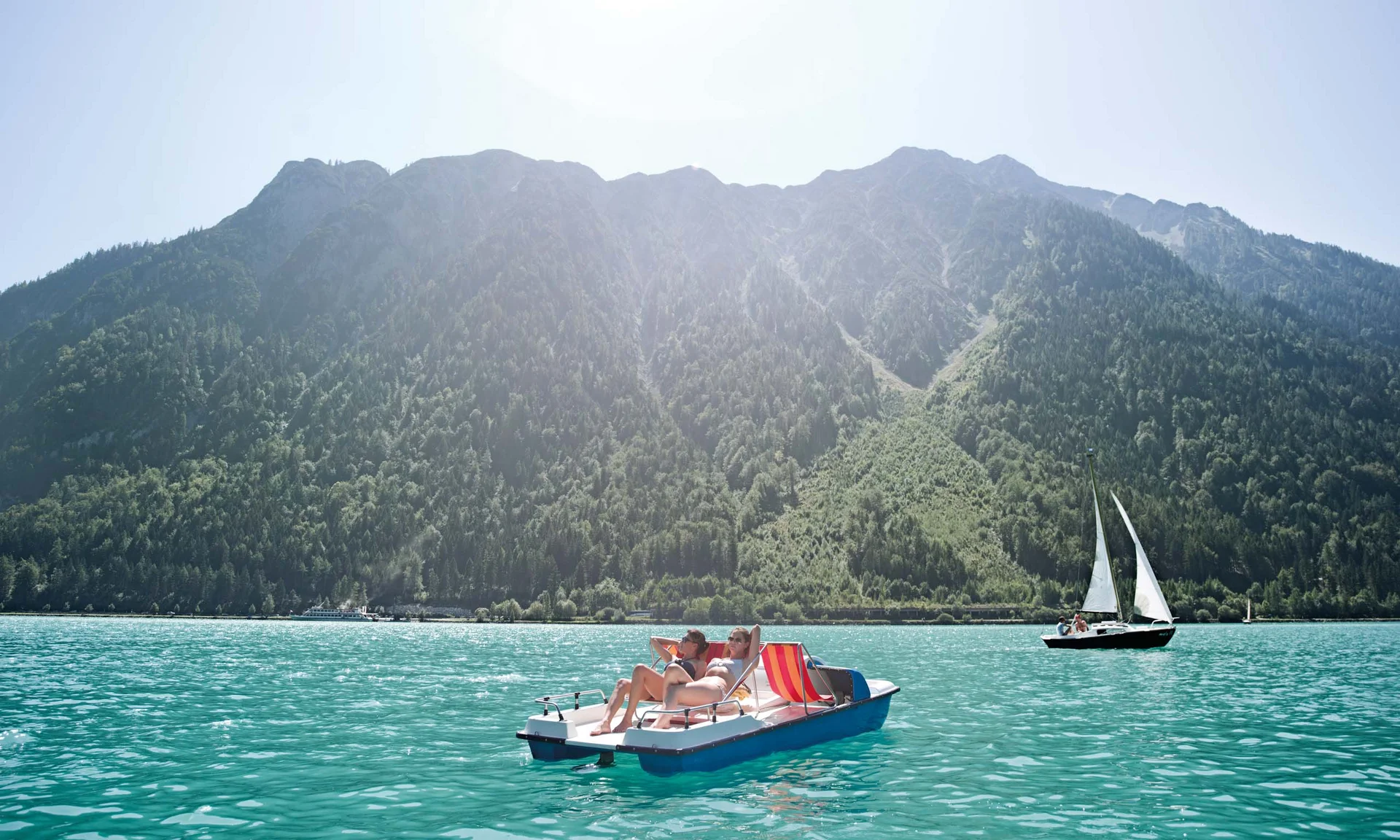 Zwei Personen sonnen sich auf einem Tretboot auf einem See vor bewaldeten Bergen