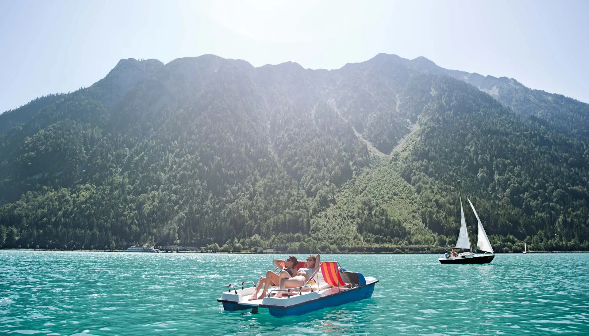 Zwei Personen sonnen sich auf einem Tretboot auf einem See vor bewaldeten Bergen
