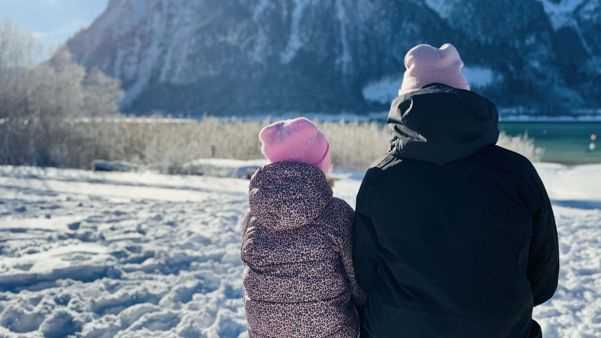 Zwei Personen mit Mützen sitzen im Schnee und blicken auf verschneite Berge