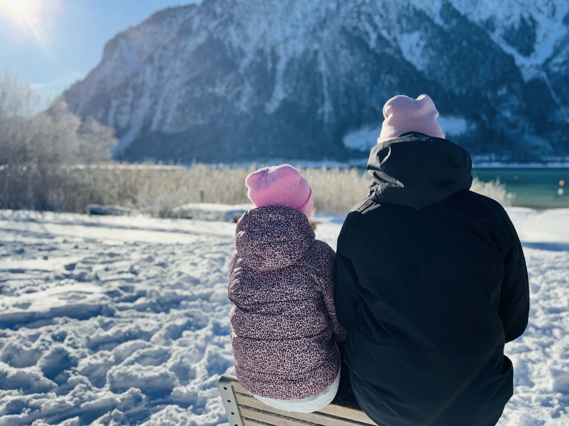 Zwei Personen mit Mützen sitzen im Schnee und blicken auf verschneite Berge