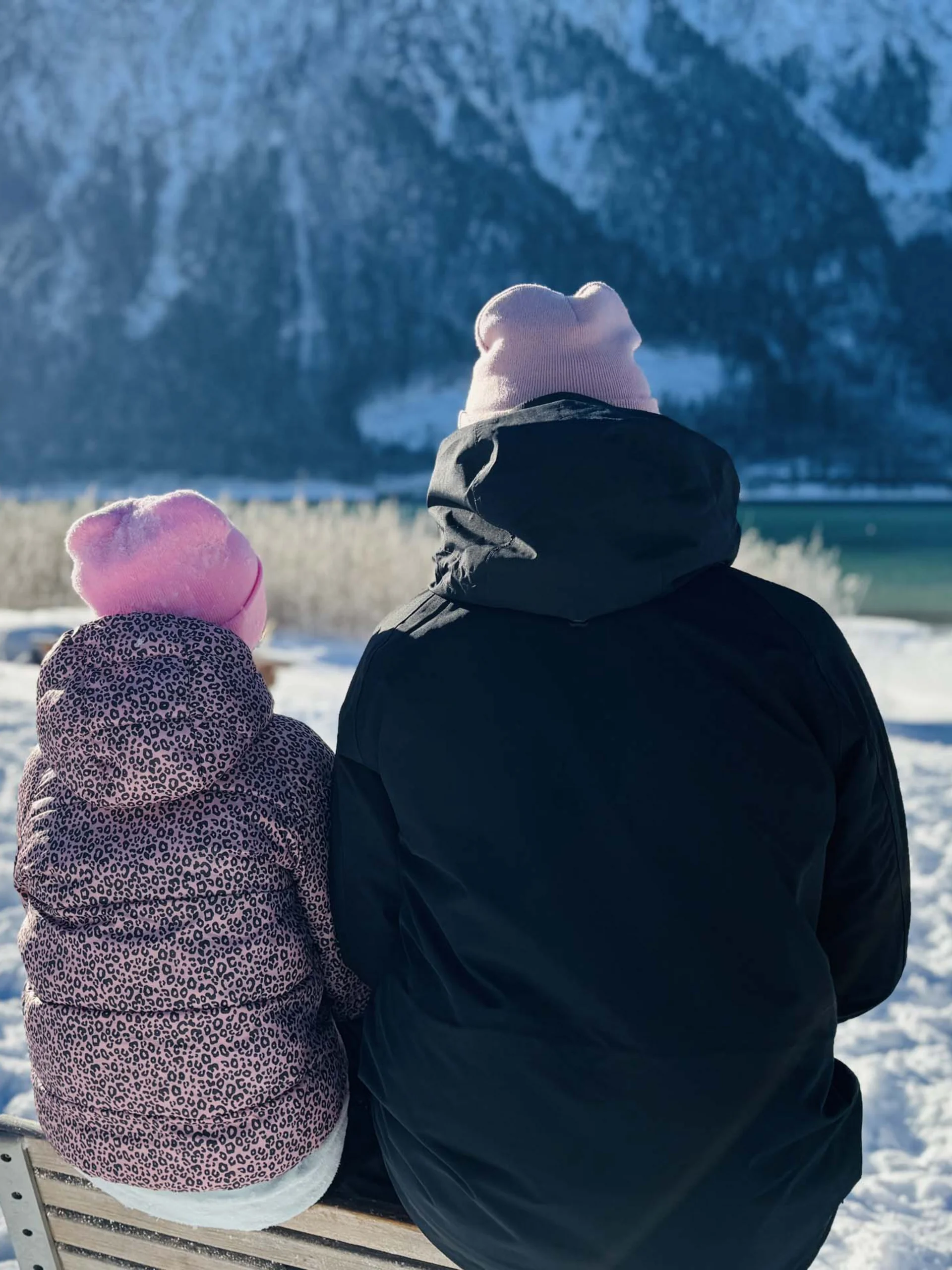 Zwei Personen mit Mützen sitzen im Schnee und blicken auf verschneite Berge