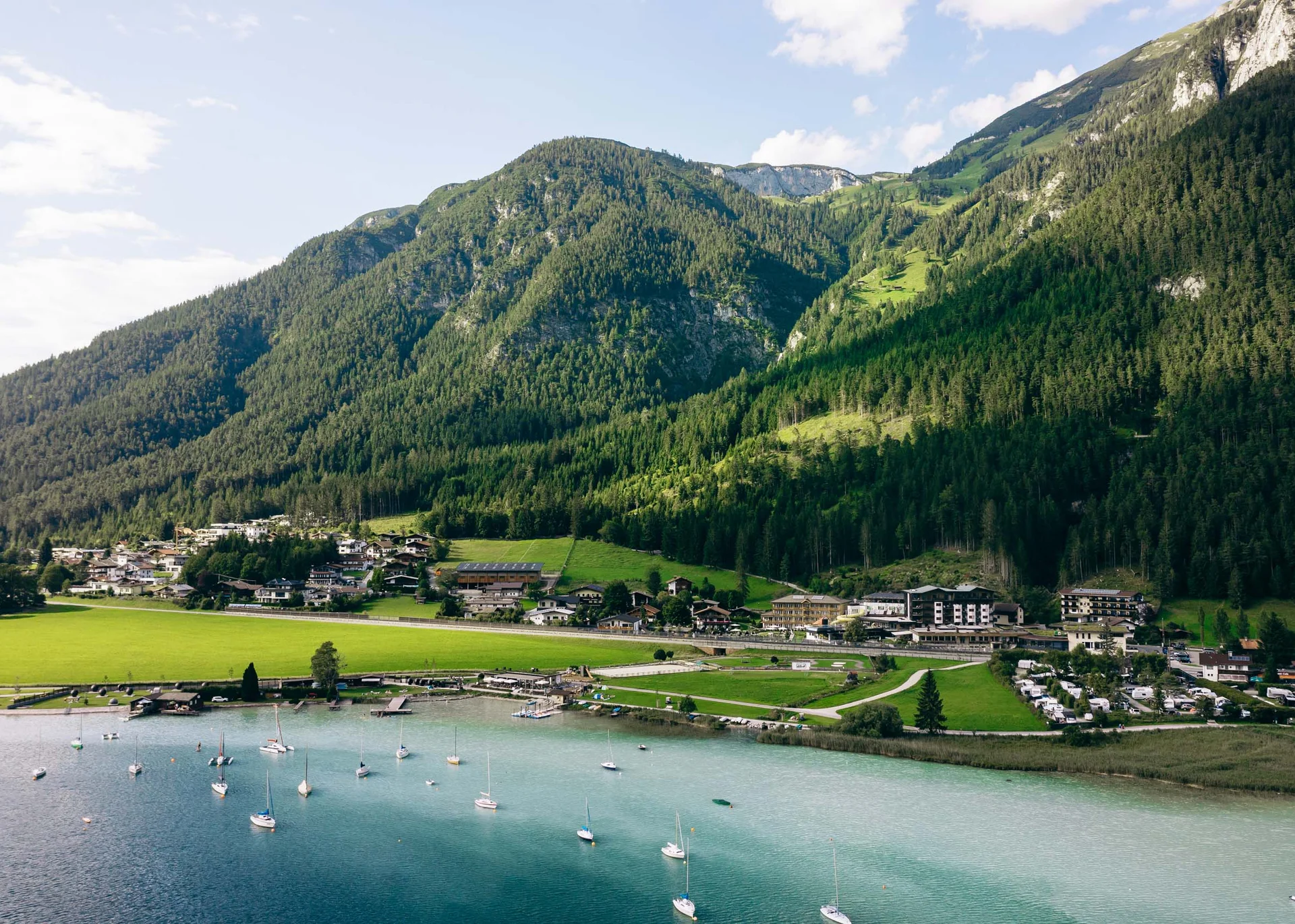 Dorf am See mit Segelbooten und bewaldeten Bergen im Hintergrund