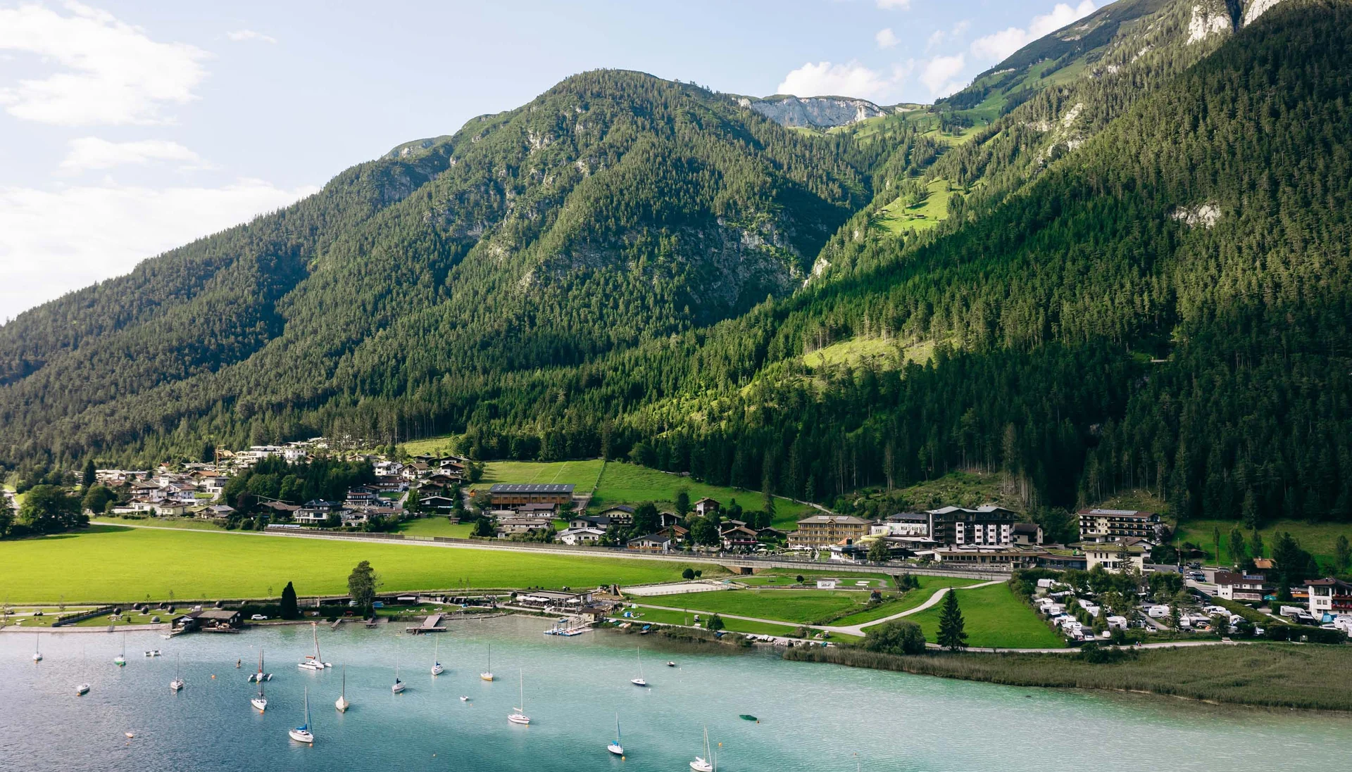 Dorf am See mit Segelbooten und bewaldeten Bergen im Hintergrund