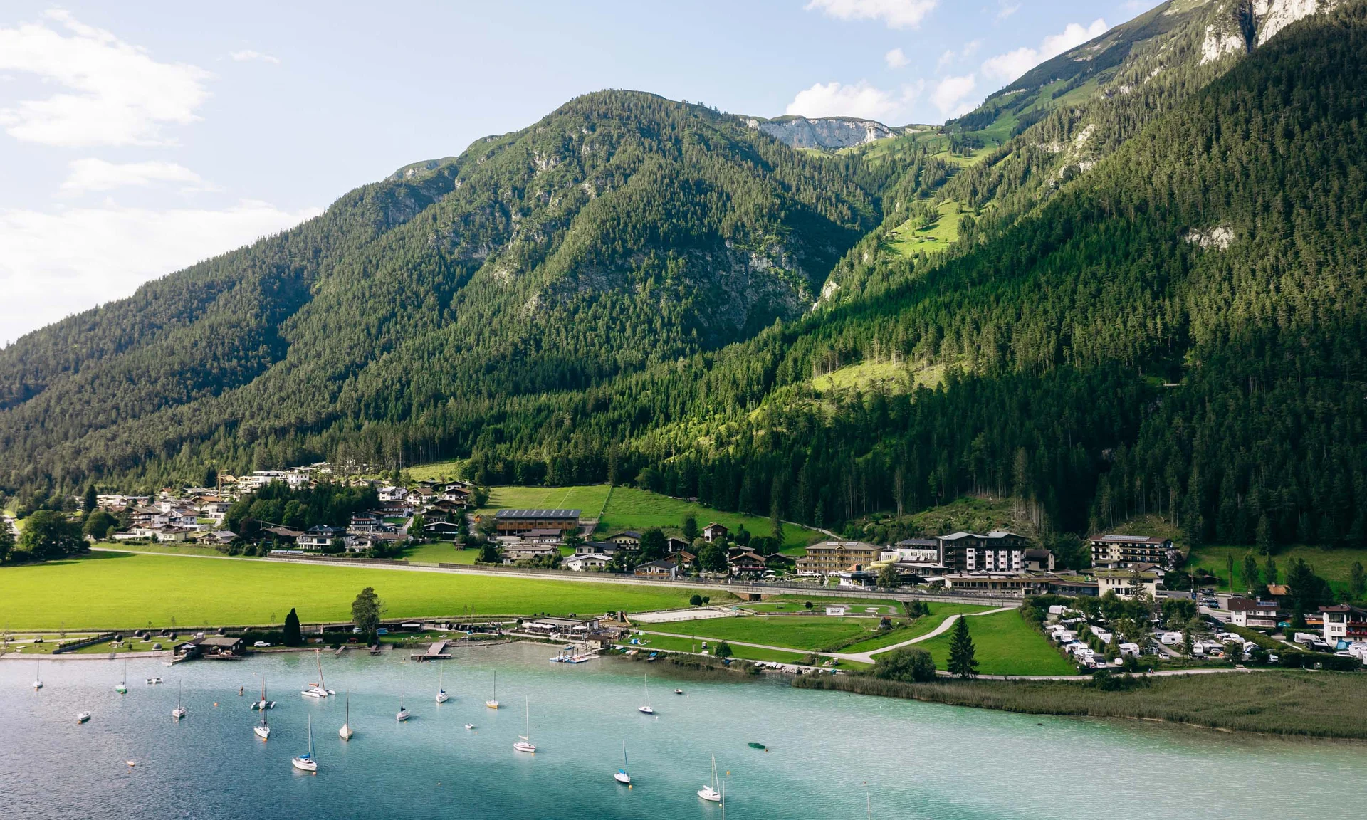 Dorf am See mit Segelbooten und bewaldeten Bergen im Hintergrund