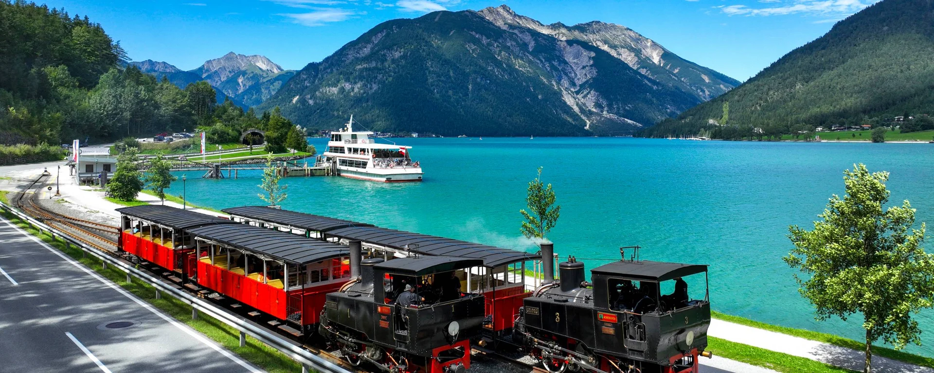 Dampflokomotive und Passagierwagen am See mit Bergen im Hintergrund
