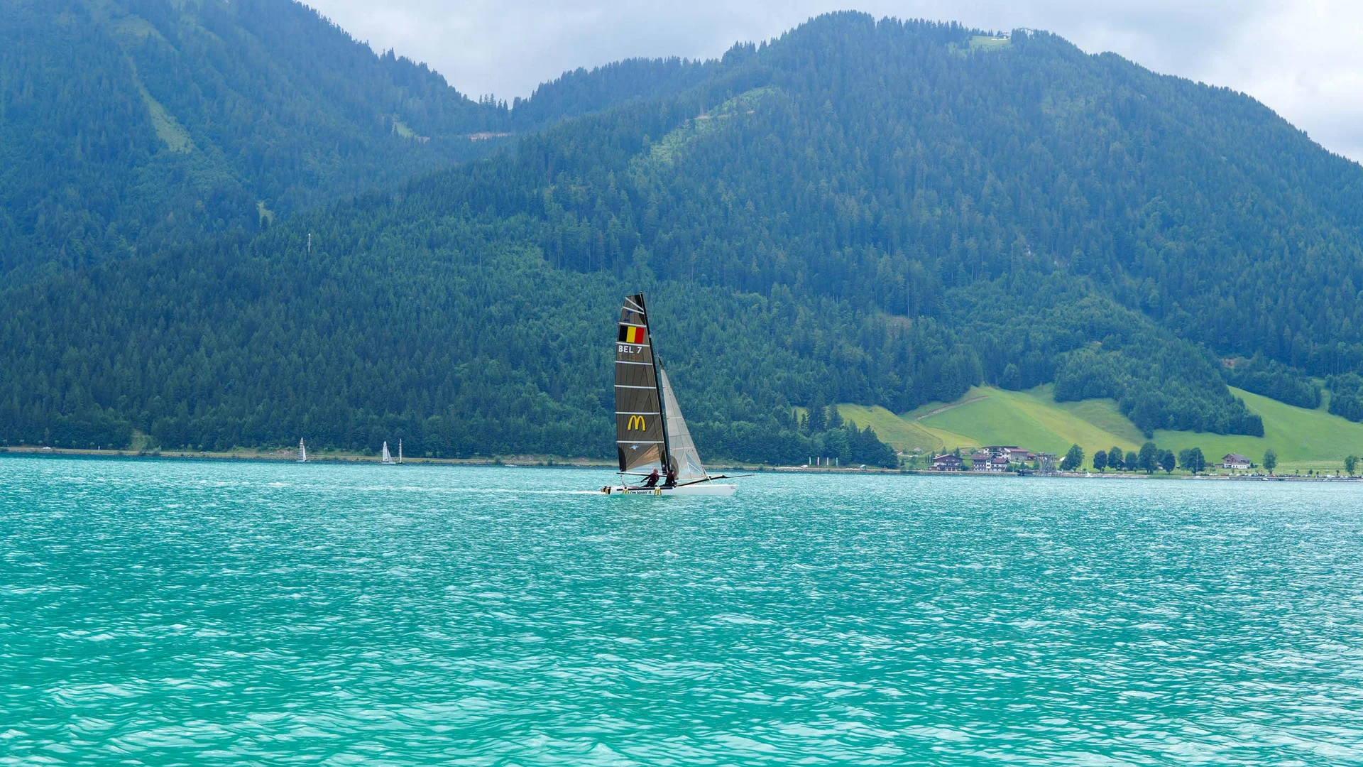 Segelboot auf türkisfarbenem See vor bewaldeten Bergen