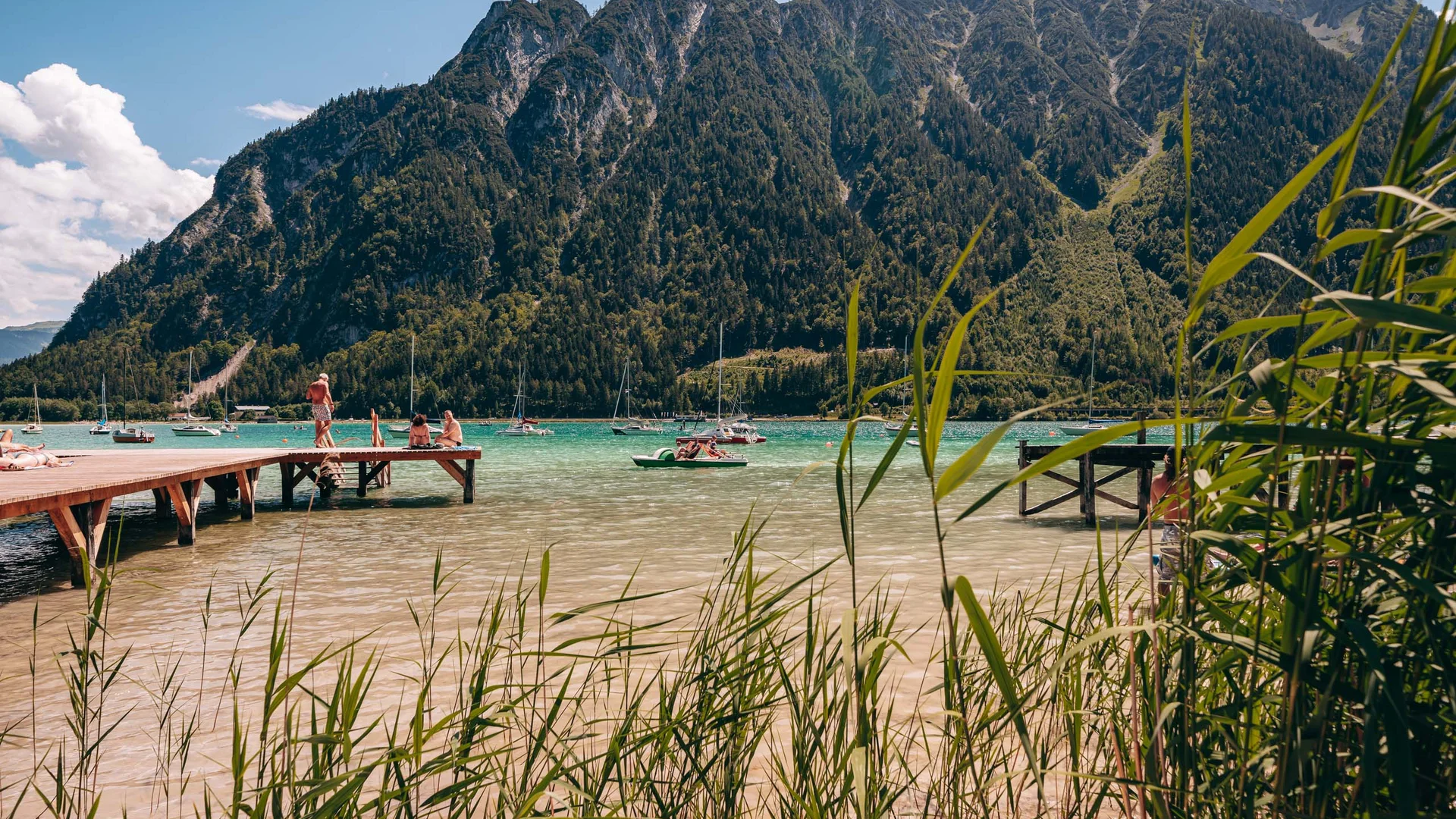 See mit Stegen, Segelbooten und Bergen im Hintergrund bei sonnigem Wetter
