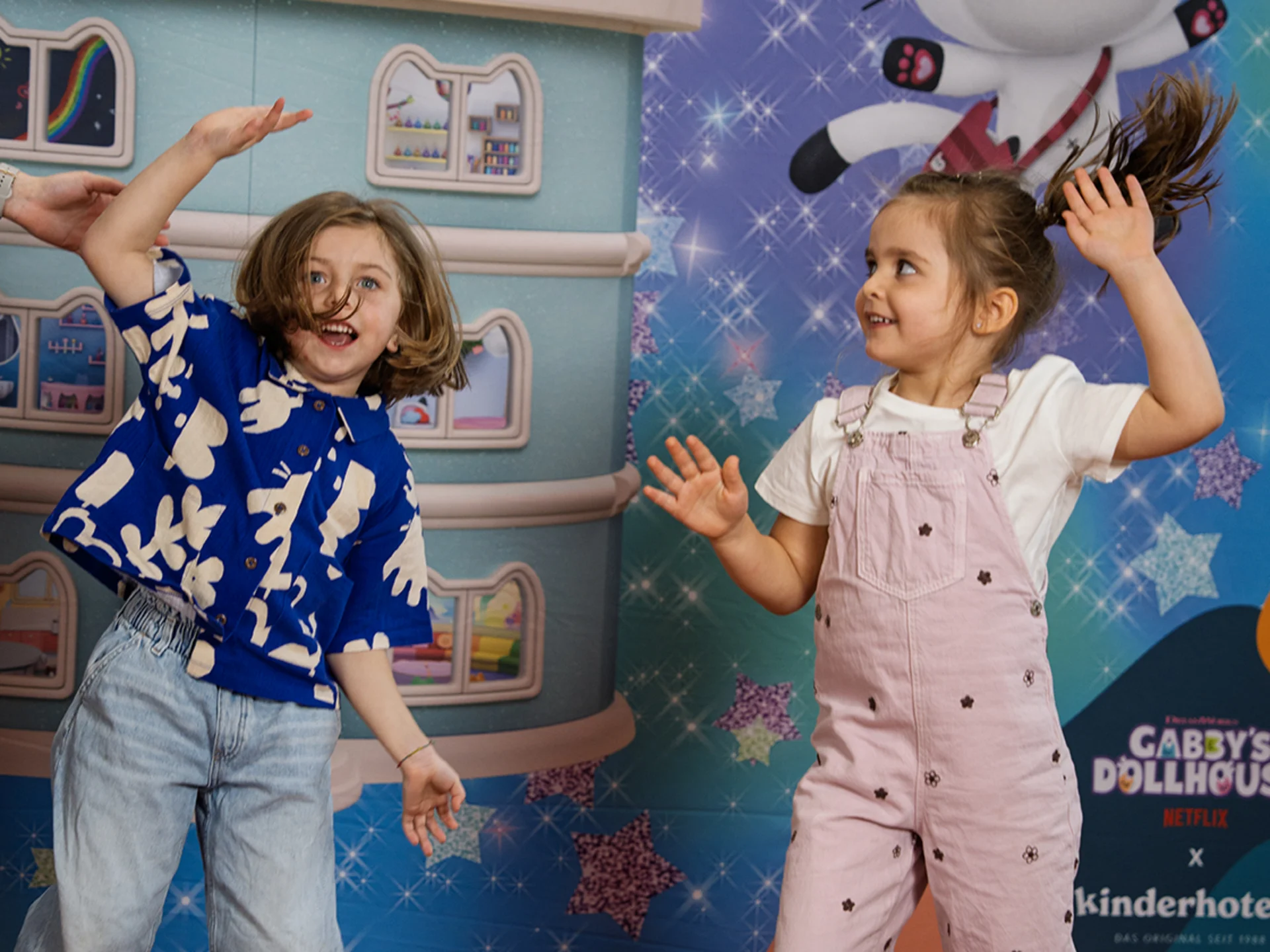 Three days full of phantasy, fun and shared happiness Two children dancing in front of a colorful Gabby's Dollhouse backdrop with toys on the floor