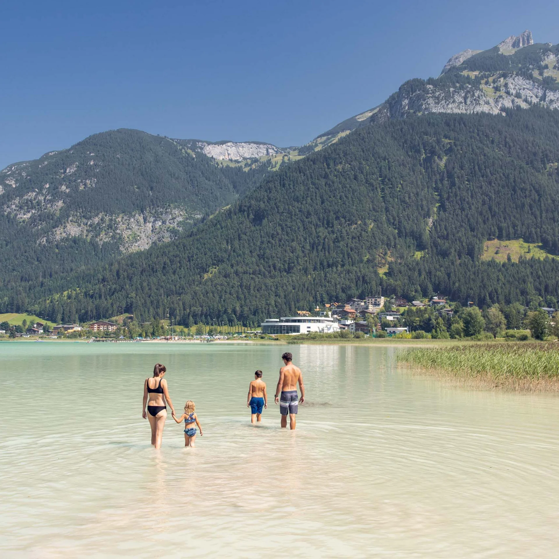 Familie watet in klarem Bergsee mit Bergen und Dorf im Hintergrund