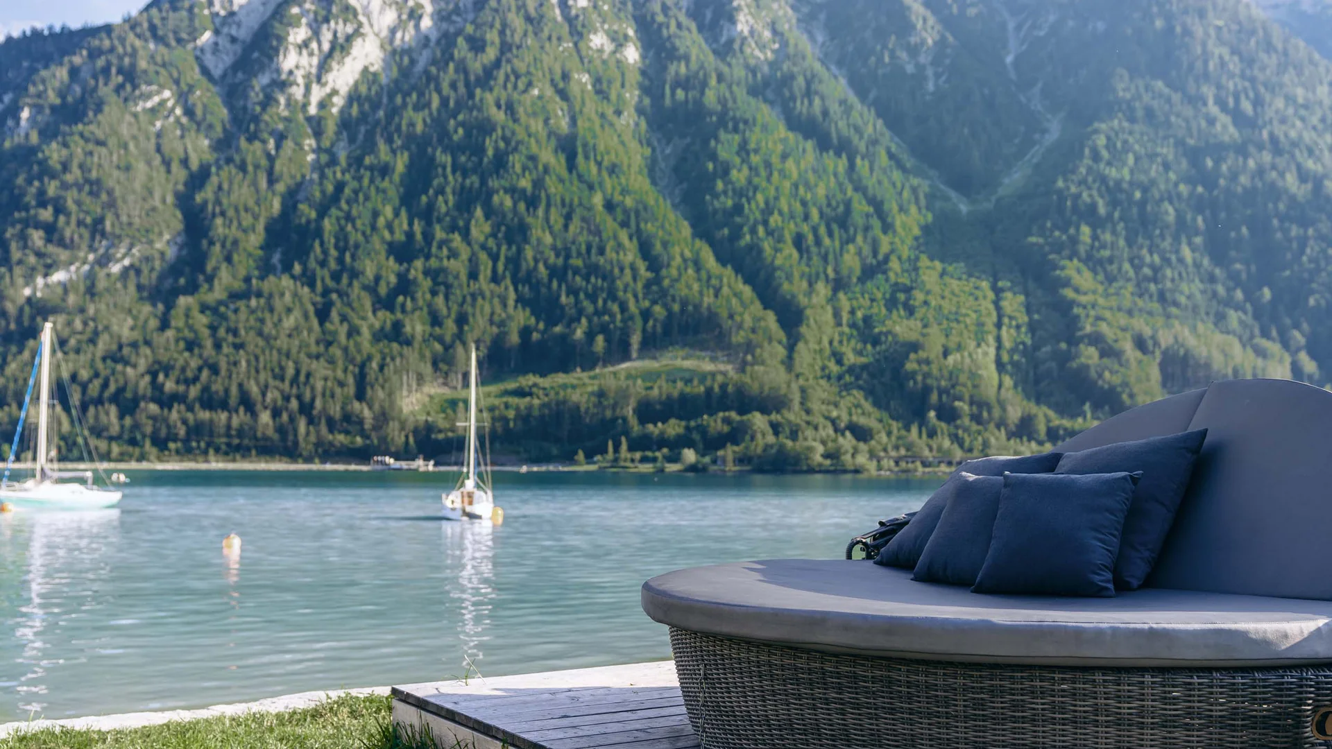 Rundes Lounge-Sofa mit Kissen am See mit Segelbooten und bewaldeten Bergen im Hintergrund