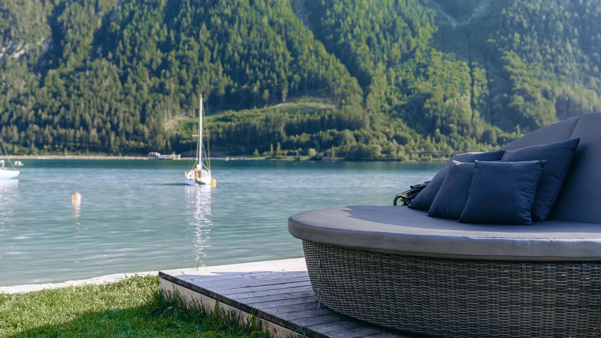 Rundes Lounge-Sofa mit Kissen am See mit Segelbooten und bewaldeten Bergen im Hintergrund
