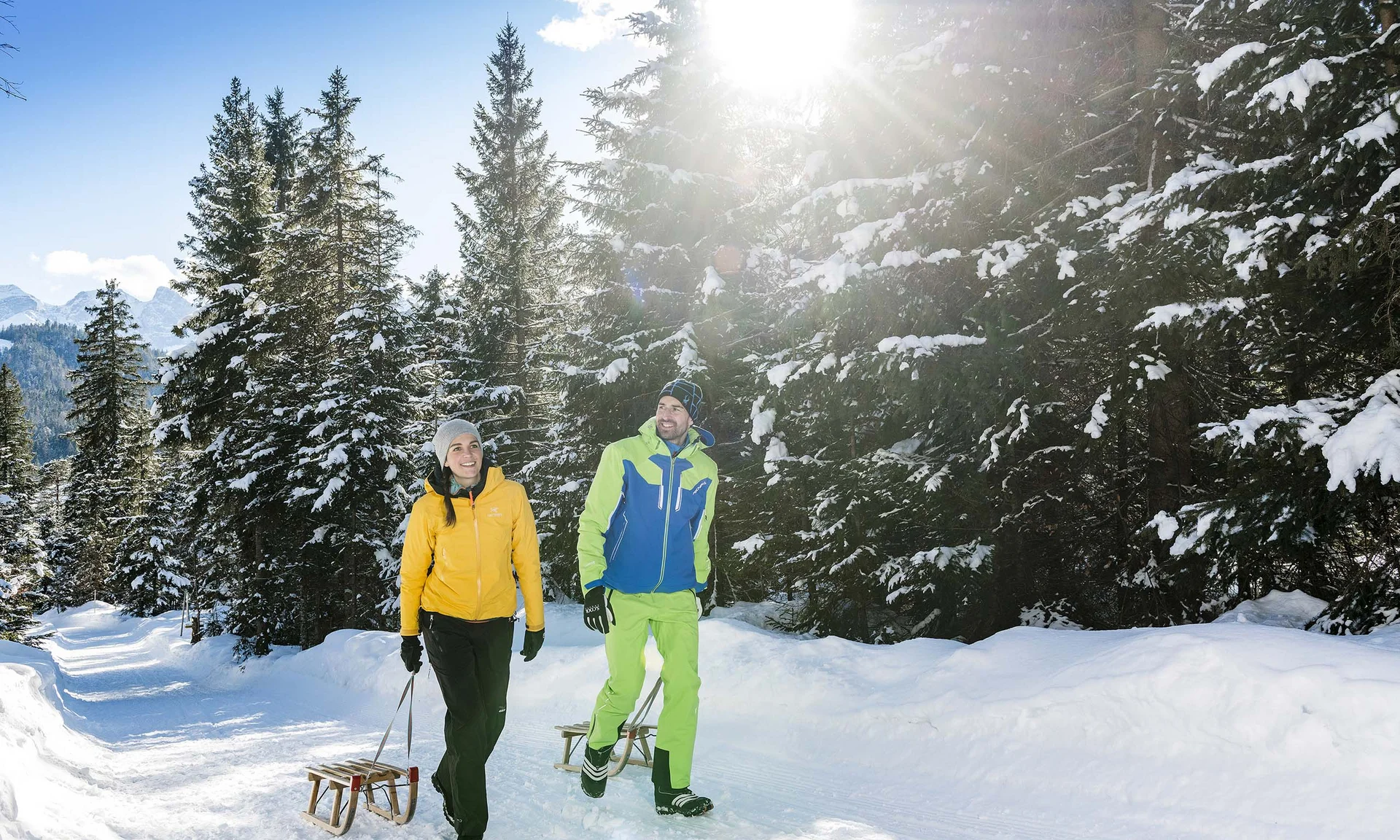 Paar spaziert mit Schlitten auf schneebedecktem Waldweg bei Sonnenlicht