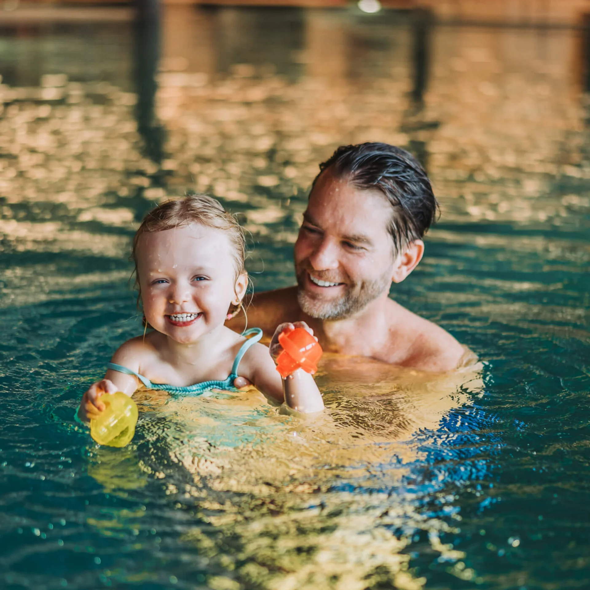 Smiling toddler with father swimming in pool holding colorful toys